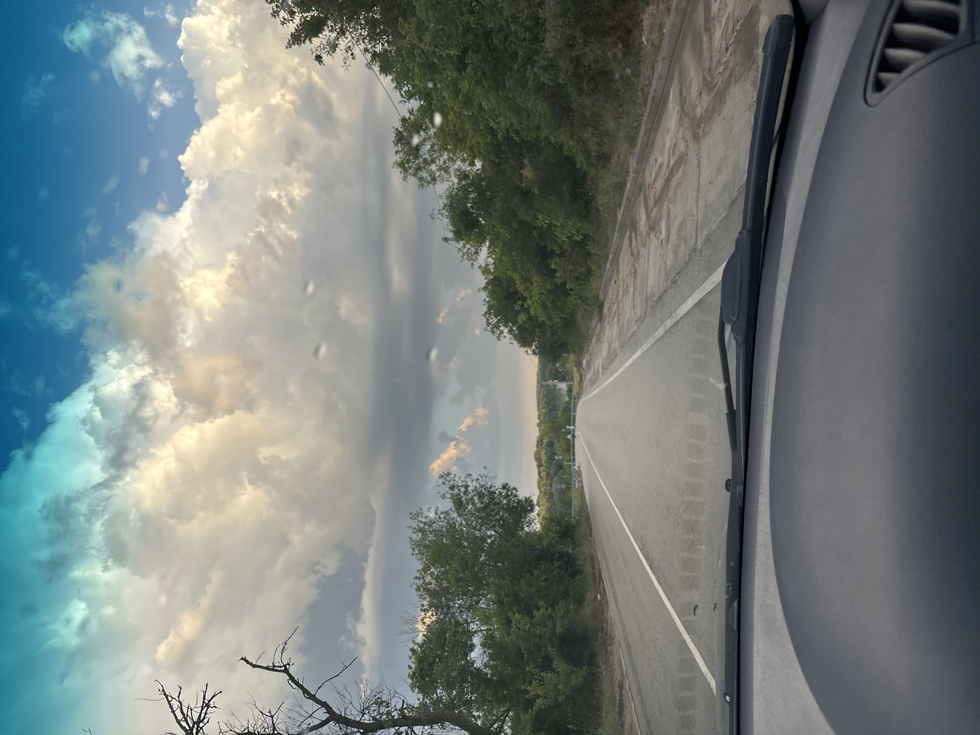 This media file captures a view from inside a vehicle, looking out through the windshield at a highway lined with trees and a concrete barrier. The sky dominates the left side of the frame, showcasing a dramatic display of clouds, ranging from bright white and yellow to dark gray, suggesting an impending storm or recent rainfall. The blue sky peeks through in some areas, creating a striking contrast.
The road ahead appears to be a two-lane highway with a white dividing line, curving slightly into the distance. The right side of the car's interior, including a portion of the dashboard and windshield wiper, is visible in the foreground on the right. Scattered droplets on the windshield are noticeable, indicating recent rain or moisture.
The scene is bathed in natural light, with the bright clouds suggesting it is either late afternoon or early morning, with the sun's rays breaking through. The presence of lush green trees along the roadside points to a temperate climate, likely during the warmer months. The concrete barrier and the road itself are typical of a rural or suburban highway setting.
The overall mood evoked by the image is one of transition and perhaps a touch of unease, given the stormy sky. The vastness of the sky and the receding road suggest a journey in progress. No people are visible, and no text is present in the image. Given the provided location context of Ladozhskaya, Russia, this scene likely depicts a road within that region, possibly on the outskirts of a populated area, given the natural elements.