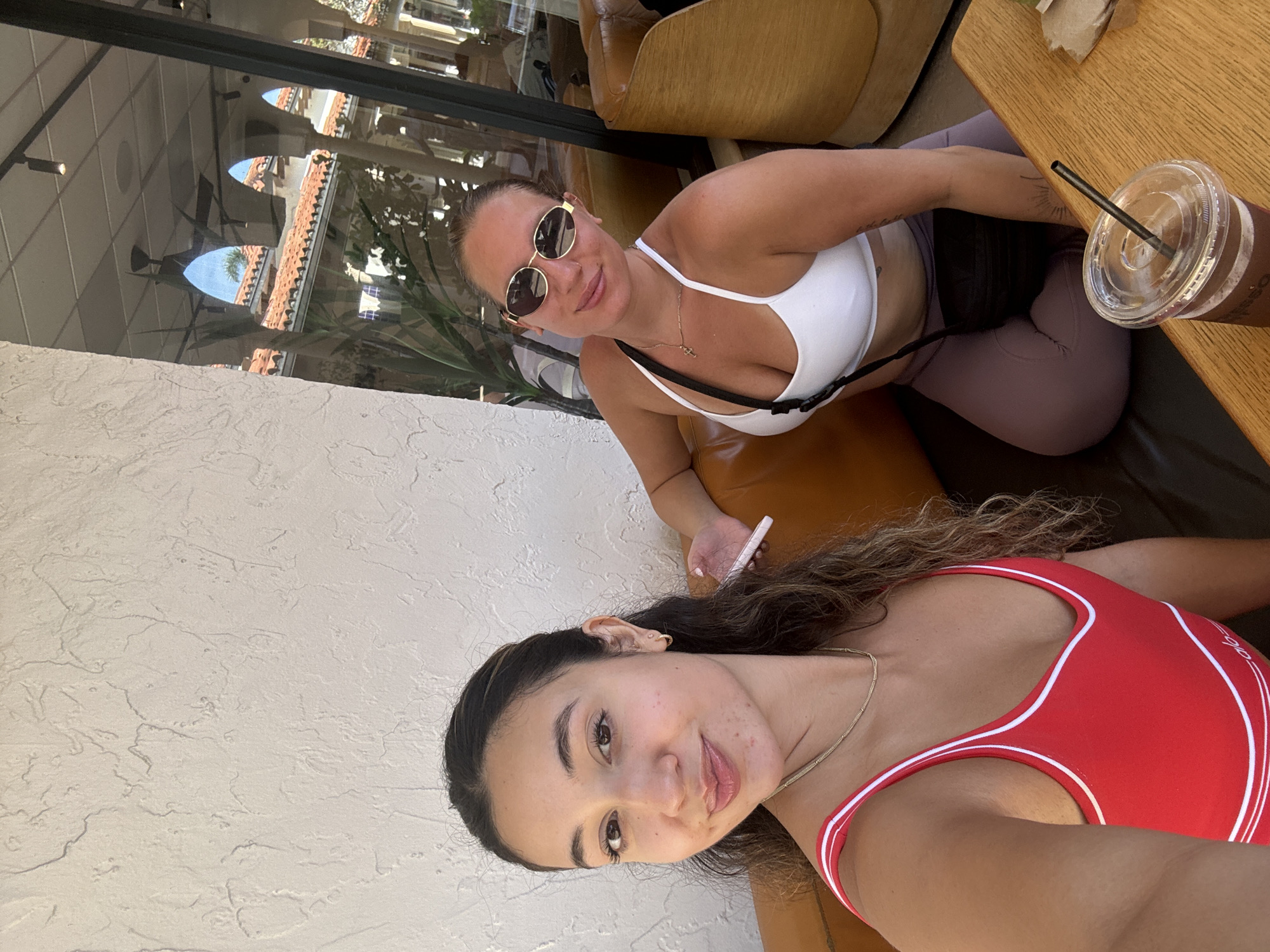In Boca Raton, Florida, United States of America, two women are seated in a cafe. The woman in the foreground, on the left, has long, dark, curly hair and is wearing a red sports bra with white trim. She is looking at the camera and smiling. The woman in the background, on the right, has her hair pulled back and is wearing sunglasses, a white sports bra, and purple leggings. She is also looking at the camera and smiling. A drink with a straw is on the table in front of them. The cafe appears to have a stucco wall and large windows, with a glimpse of Mediterranean-style architecture outside.