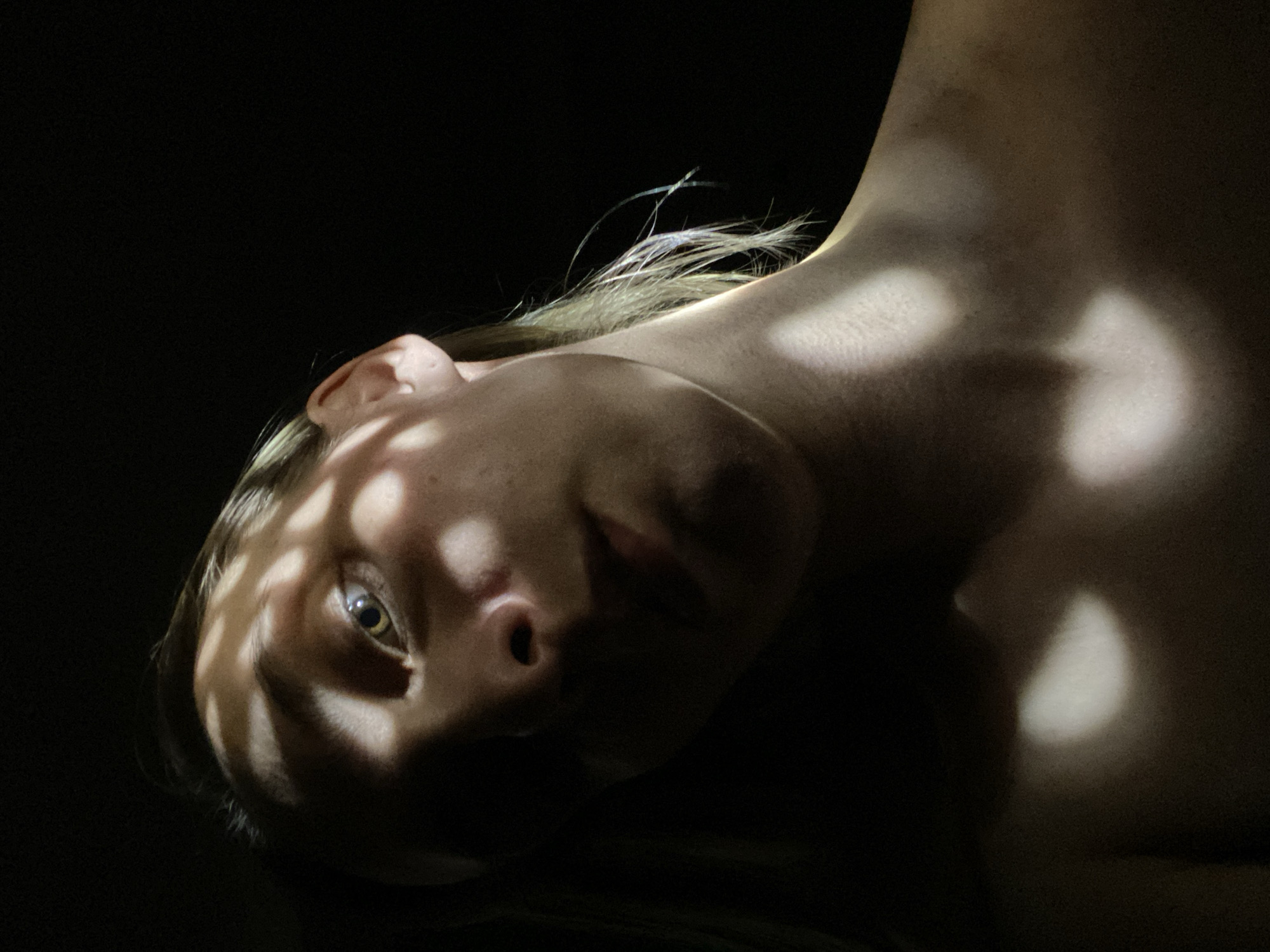 The image shows a close-up of a person's face and upper torso, likely female, against a dark background. The person is lying down, tilted towards the viewer. The skin is fair, with a pattern of light and shadow creating a latticework effect across the face. One eye is clearly visible and appears to be hazel or green. Light shines on the neck and shoulder. The location in the city of Chaikovsky, Russia is not discernible from the image.