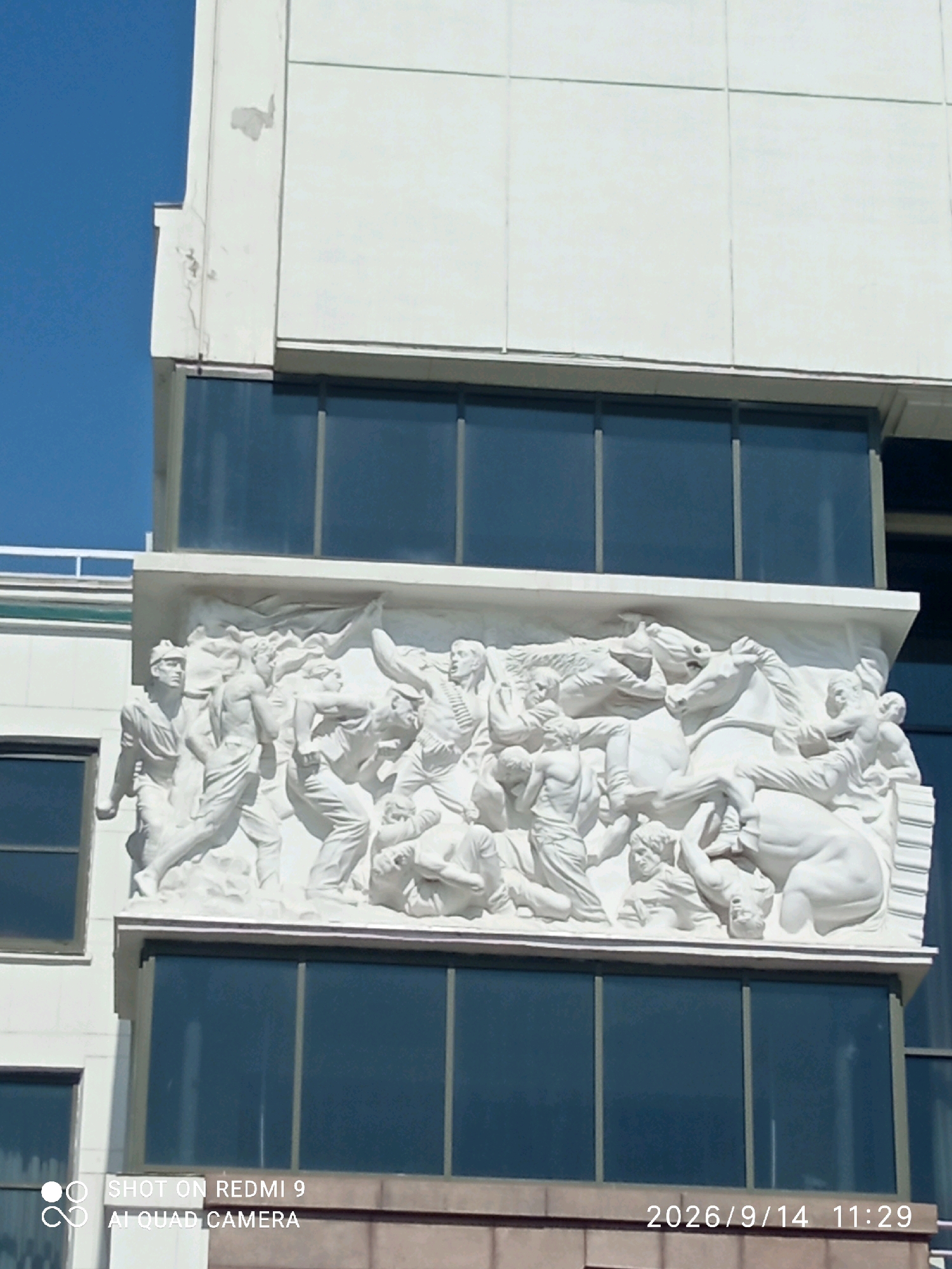 This image shows a white bas-relief sculpture depicting a chaotic scene of people and horses, mounted on the facade of a modern building with dark glass windows. The sculpture appears to be a historical or allegorical representation, with figures in dynamic poses, some falling, others struggling, and horses in motion. The building itself is contemporary, with clean lines and large windows reflecting the sky. At the bottom right of the image, a timestamp indicates "2026/9/14 11:29". Below the timestamp and on the left side, there is a watermark stating "SHOT ON REDMI 9 AI QUAD CAMERA". The sculpture is located on the facade of a building in Rostov-on-Don, Russia. The bas-relief is positioned above the second floor of windows and spans a considerable width. The overall scene is illuminated by daylight, creating distinct shadows that add depth to the sculpted figures.