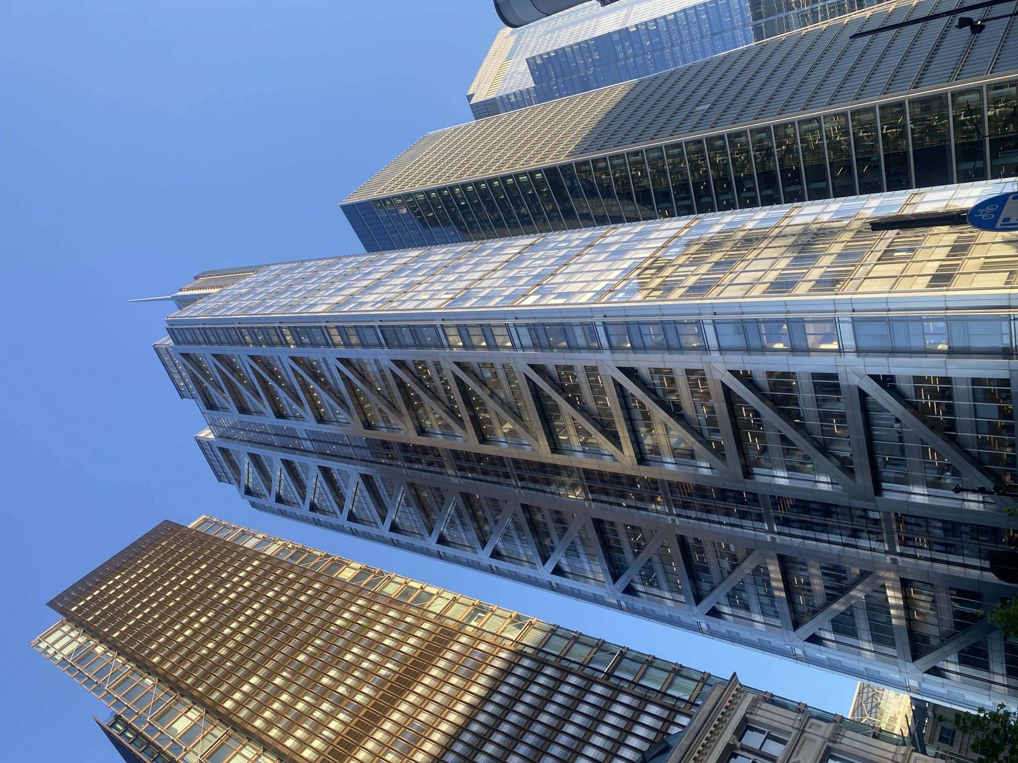 This low-angle, upward-looking shot captures several modern skyscrapers against a clear, bright blue sky in London, United Kingdom. The perspective emphasizes the towering height and architectural details of the buildings.
The most prominent structure, located centrally, is a very tall building characterized by a distinctive external diagonal lattice or structural bracing system. This building features extensive glass facades reflecting the sky and surrounding environment. Its upper sections have a more uniform glass and metal finish, and a slender spire or antenna is visible at its apex against the empty sky in one of the crops.
To the left of the central building, a slightly shorter, box-like skyscraper is visible. This building features a facade composed of numerous square or rectangular panels, appearing metallic or tinted glass, reflecting warm sunlight. In the lower part of the image, the base of this building appears to integrate with an older, more traditional stone or masonry building with classic architectural details.
Further behind the main central building, additional skyscrapers with reflective glass surfaces are partially visible, suggesting a dense urban environment. The sunlight is bright, indicating a sunny day, likely morning or late afternoon, as suggested by the warm reflections on some building surfaces and the lack of harsh, overhead shadows. The sky is completely cloudless.
No people are visible. The primary activity is the architectural presence of these modern structures in a bustling city. A small, blue circular sign with a white bicycle symbol, likely indicating a cycling amenity, is partially visible on one of the buildings' lower sections. The overall impression is one of modern urban development and impressive scale.