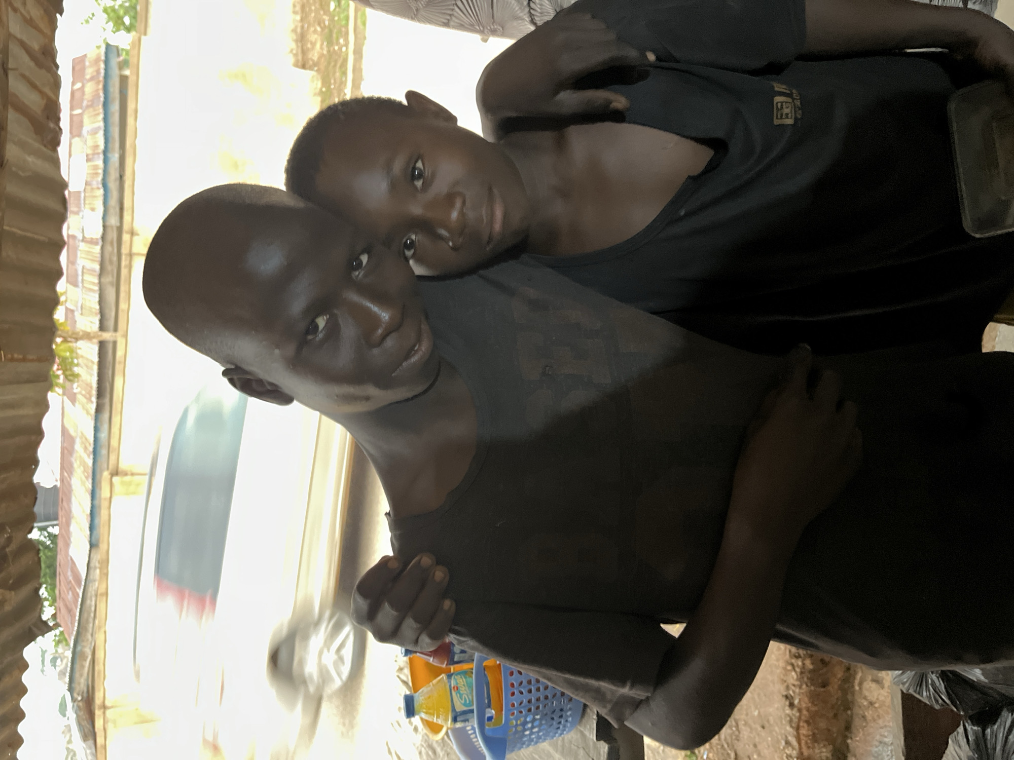 Two dark-skinned individuals are visible, one with a shaved head. Both are wearing dark clothing. They appear to be in a hug or embrace. The location is Jos, Nigeria. A vehicle is seen in motion in the background. In the foreground, there is a blue plastic basket and bottles of yellow liquid. The environment includes corrugated metal structures.