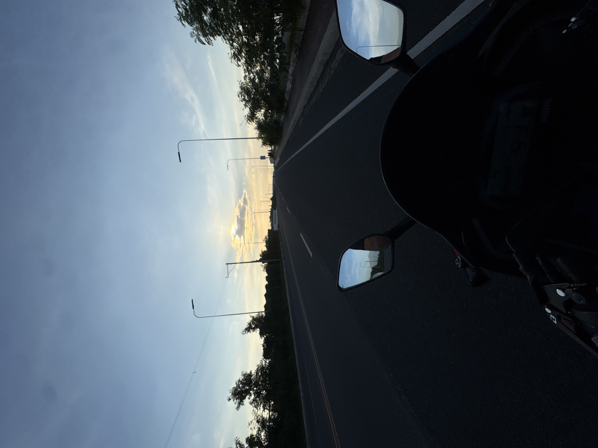 The image captures a perspective from a motorcycle traveling on a multi-lane road during twilight. The sky displays a gradient of blue and yellow hues, with scattered clouds and the sun setting on the horizon, casting a warm glow. Along the sides of the road, there are trees and vegetation, with streetlights lining the route. In the foreground, the motorcycle's windshield and mirrors are visible. The right mirror reflects the road and the sky, showing streetlights and trees. The left mirror also reflects the sky and streetlights. The road itself has white lane markings. The overall scene suggests a peaceful journey along an urban or suburban highway in Davao City, Philippines, as the day transitions into night.