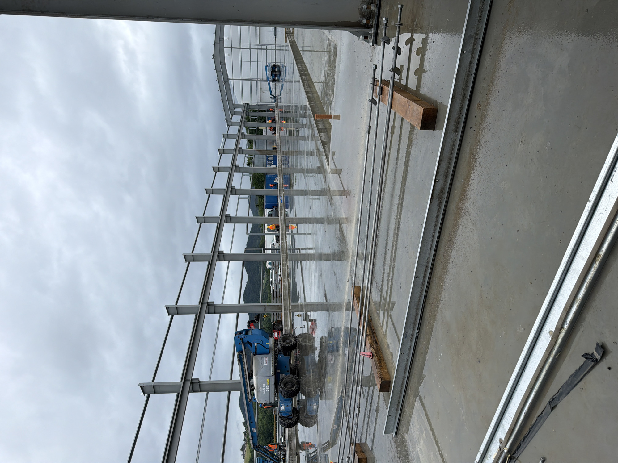 An exterior, eye-level, wide shot captures an overcast day at a construction site in Janowice, Poland. The foreground of the image is dominated by a wet concrete floor, with visible reflections of the surrounding structures and sky. Several horizontal metal beams, some with nuts and bolts, are laid out on the floor, supported by wooden blocks in places.
In the mid-ground, a blue Genie brand aerial work platform is parked on the wet surface. The machine displays the model number Z-80/60. Several construction workers, wearing orange vests, are visible in the mid-ground, some walking and one standing near the aerial platform. Further back, the skeletal structure of a building is under construction, with large metal columns and beams forming its framework. A partially constructed roof structure is visible towards the right side of the image. In the background, rolling green hills with trees are visible under a cloudy sky. Other construction equipment and vehicles, including a shipping container, are also present in the mid-ground and background. The overall scene depicts an active construction project on a damp day.