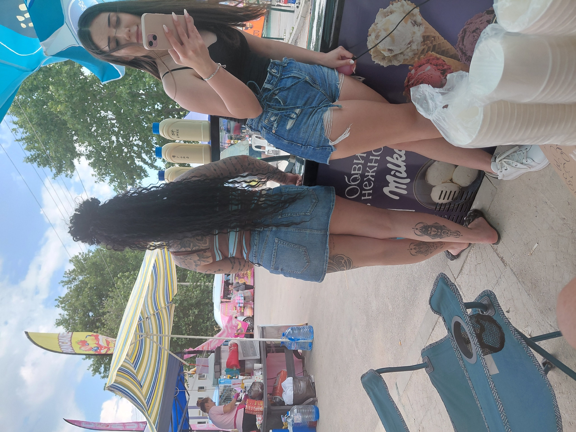 The image captures an outdoor market or fair scene on a sunny day with a blue sky and scattered clouds. In the foreground, two women are positioned near a vendor counter. The woman on the left, viewed from behind, has long, dark, curly hair gathered into a bun. She wears a light blue strapless top and a denim mini-skirt. Visible tattoos adorn her upper back, right arm, and both calves, depicting stylized patterns. She is wearing black flip-flops.
To her right, a second woman, seen in profile, has long dark hair and wears a black tank top and ripped denim shorts, complemented by white sneakers. She holds a light-colored smartphone in both hands, looking down at the screen. A silver bracelet is visible on her left wrist. On the counter in front of them are several white plastic bottles with blue caps, bearing Cyrillic text, possibly containing syrups or condiments. Stacks of white paper cups are also visible on a display featuring "Milka" branding and images of ice cream cones.
Further into the midground on the left, a vendor stall is covered by a yellow and blue striped umbrella. A woman in a pink shirt is working behind the stall, which has a red dispenser and various items on its table. Two large blue water jugs rest on the ground beside the stall. A tall yellow flag with "PANCAKE" written vertically and an image of pancakes with fruit and cream is prominent above this stall.
The background consists primarily of dense green trees under the clear sky, with overhead utility wires stretching across the upper portion of the frame. On the far right, a pink food truck with Cyrillic text is visible, with a person standing near it. The ground surface is paved, suggesting a public outdoor area in the city of Unknown, Unknown.