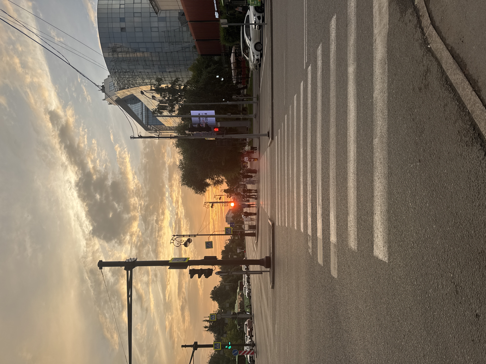 The image captures a sunset over a city intersection in Tyumen, Russia. A crosswalk dominates the foreground, with several lanes of traffic extending into the distance. Cars and buses are visible on the road, and pedestrians are crossing the street. A modern glass building with scaffolding on one side stands to the right of the intersection. The sky is filled with dramatic clouds illuminated by the setting sun. Traffic lights and street lamps are also present, indicating a typical urban environment. The scene appears to be a busy street during evening rush hour.