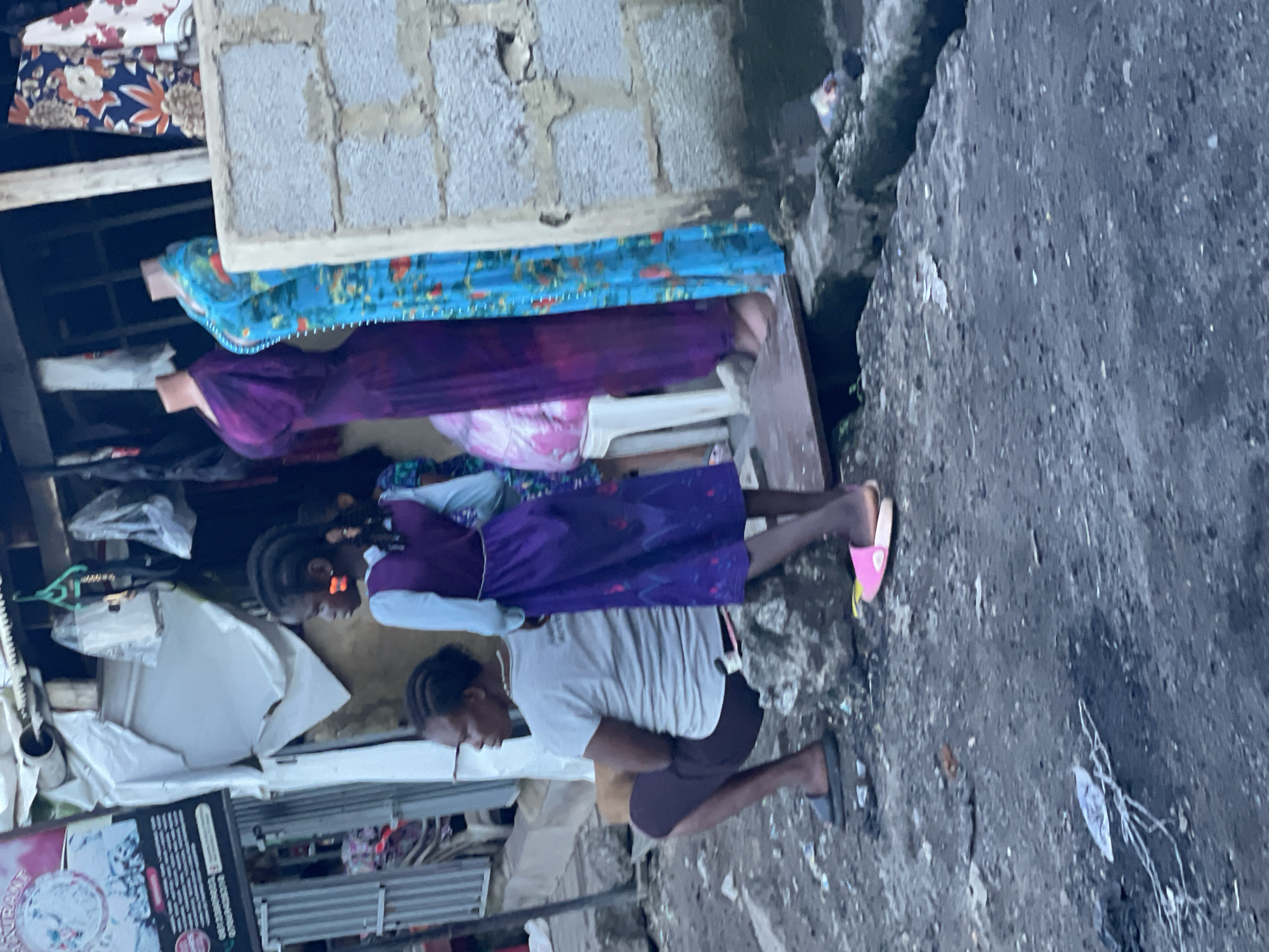 The image depicts a street scene in Abuja, Nigeria, showing a modest outdoor market or stall. In the foreground, a person is seated on rocks, facing left, wearing a light-colored t-shirt and dark trousers, with black slippers on their feet. To their right, a young girl stands, facing left, wearing a purple dress with a white collar and large orange earrings. Behind them, several mannequins display brightly colored dresses; one is a deep purple, and another is turquoise with a floral pattern. The background reveals a makeshift structure with corrugated metal and fabric coverings, suggesting a temporary or informal business setup. Various items, including plastic bags and clothing on hangers, are visible hanging from the structure. The ground is rough and uneven, with debris scattered about. A sign with text and an image is visible on the left side of the frame. The overall impression is one of a bustling, informal commercial area in an urban setting.
