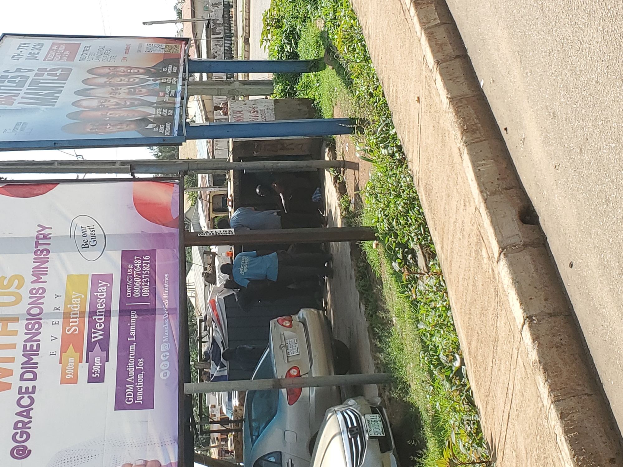 The image depicts a street scene in Bukuru, Nigeria, likely near a commercial or public gathering space. A large billboard advertising "GRACE DIMENSIONS MINISTRY" with event details for "Sunday" at 9:00 am and "Wednesday" at 5:30 pm is prominently displayed. Adjacent to it is another billboard for an event titled "BATTLES & MANTLES" scheduled for "4TH - 7TH JUNE, 2026."
In the foreground, several vehicles are parked, including a silver Toyota Corolla and a lighter-colored sedan. A group of individuals, some appearing to be ministry members or event staff, are gathered near the billboards and some structures that might be makeshift stalls or service areas. One individual in a blue t-shirt with "Micron Waves" printed on it is visible. Another person is seen sitting inside a small enclosed space.
The environment suggests a typical Nigerian urban setting with paved roads, sidewalks, and abundant greenery. The presence of utility poles and overhead wires further indicates an inhabited area. The overall atmosphere appears to be one of community activity, possibly related to religious services or local events.
