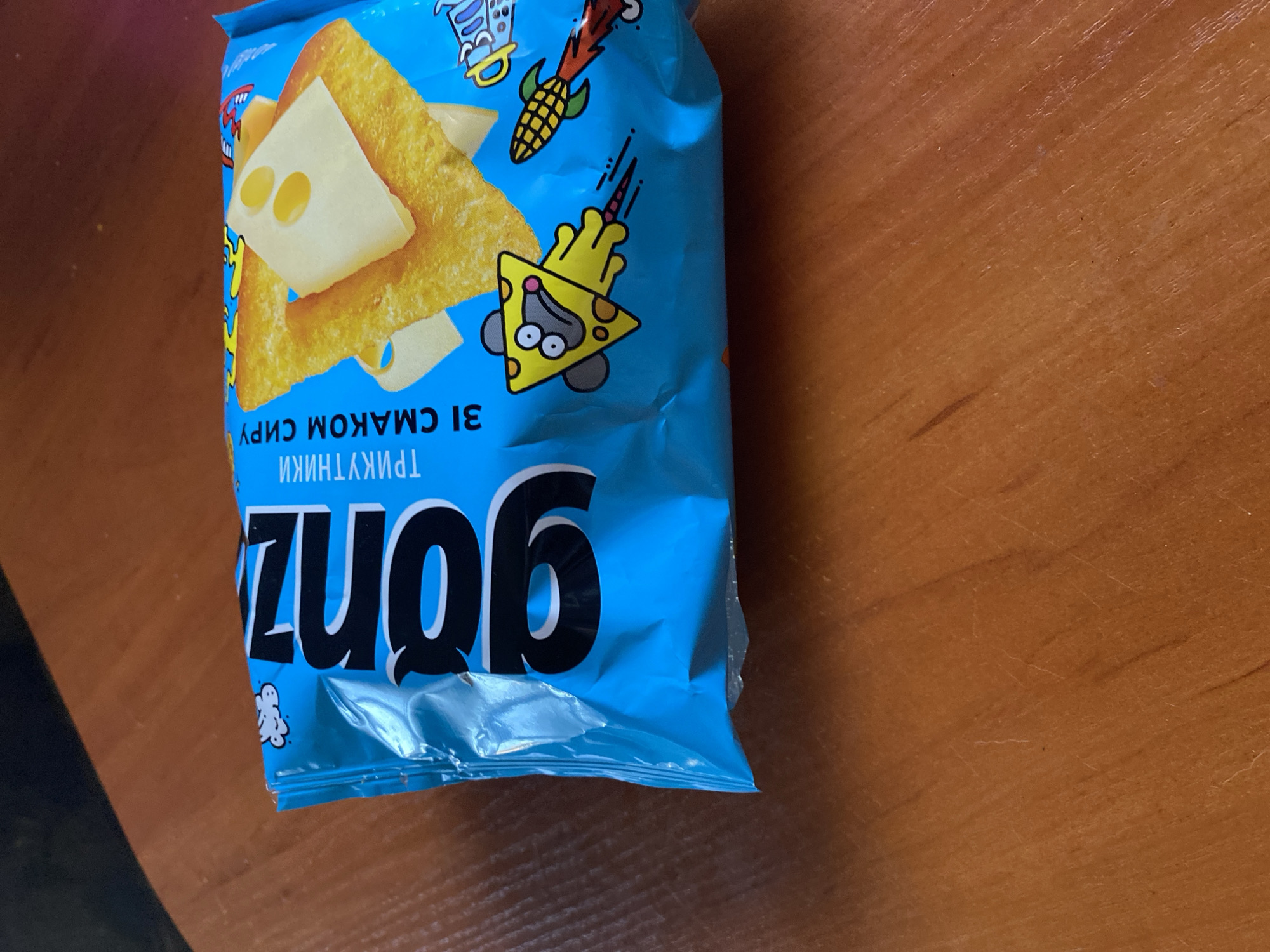 The image shows a close-up, overhead view of a bright blue snack bag lying on a wooden table. The bag features a large, bold, black logo that reads "gonz". To the right of the logo, text in Ukrainian reads "ТРИКУТНИКИ ЗІ СМАКОМ СИРУ", which translates to "TRIANGLES WITH CHEESE FLAVOR". The bag's design also includes cartoon illustrations of a mouse-shaped triangle with cheese holes, a rocket-shaped corn cob, and what appears to be a stylized rocket. Several cheese-covered triangular snacks are visible through the clear plastic window on the bag. The wooden table surface shows a visible wood grain and some minor scratches. There is no indication of the location being Nova Praha, Ukraine.