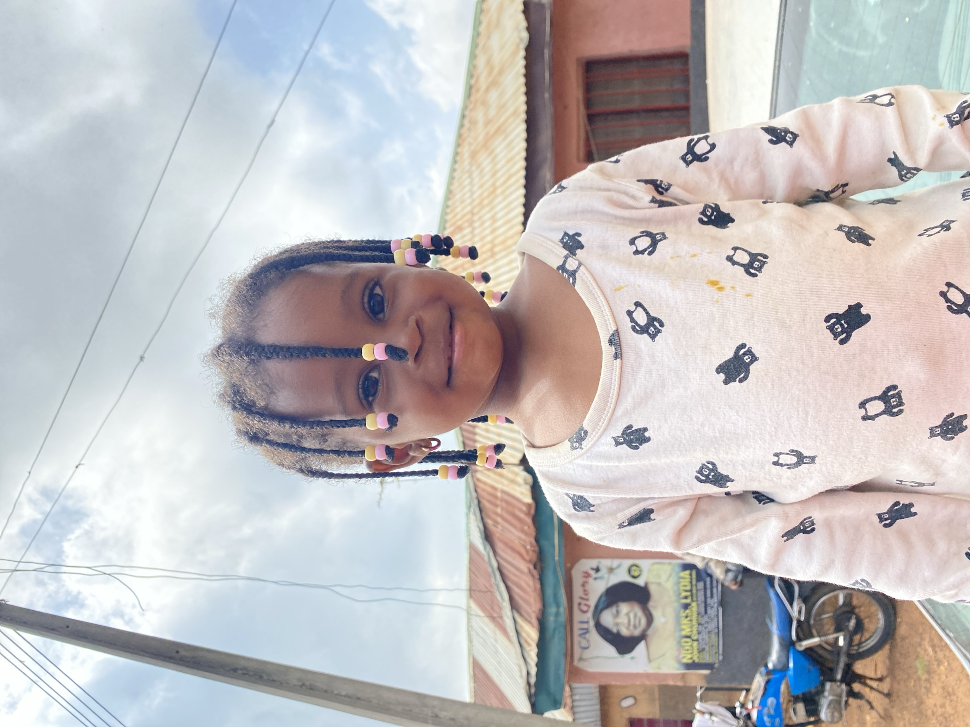In Bukuru, Nigeria, a young girl with braided hair adorned with pink and yellow beads smiles at the camera. She is wearing a white long-sleeved shirt patterned with black bear figures. Behind her, a pink building with a corrugated iron roof stands under a cloudy sky. A poster is affixed to the building, featuring a portrait of a woman with the text "CALL Glory NGO MRS. LYDIA". To the left of the building, a blue motorcycle is parked. A utility pole with overhead wires is visible on the far left of the frame. The scene appears to be an outdoor street view.