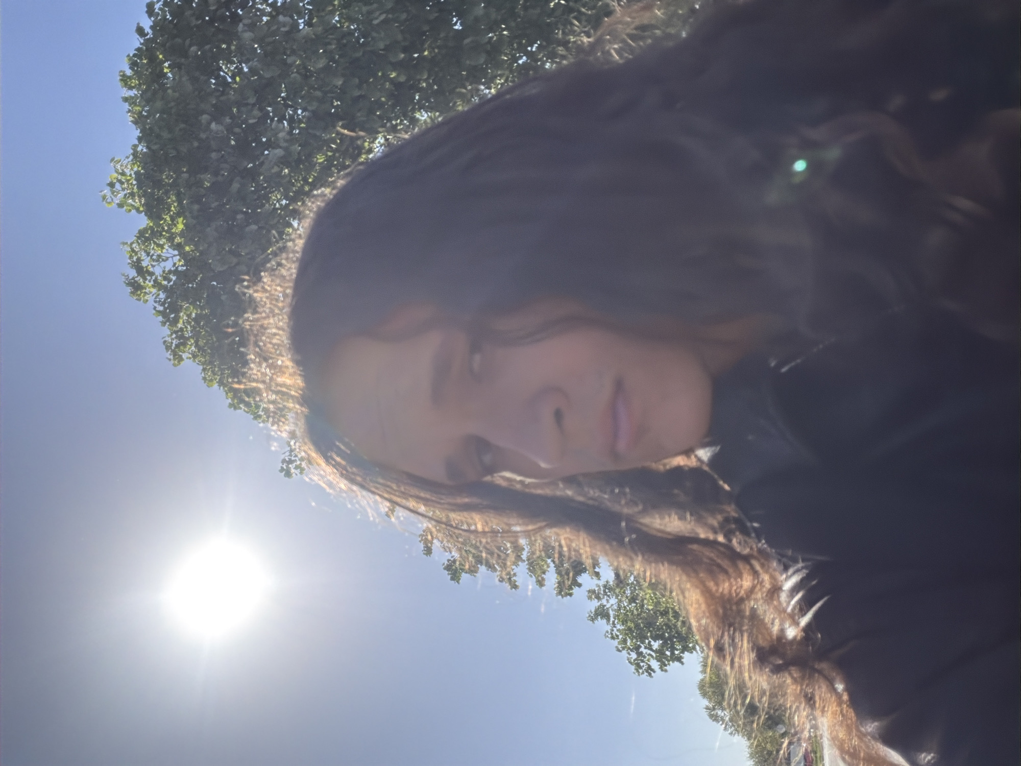 The image is a brightly lit, vertically oriented outdoor shot, rotated approximately 90 degrees counter-clockwise. The primary subject is a person with long, dark, wavy hair, seen from an upward angle. Their face is partially visible, looking towards the camera with a neutral to slightly serious expression. Their hair catches the sunlight, creating a bright halo effect around their head. They appear to be wearing a dark garment.
In the background, a clear, bright blue sky dominates the scene. A prominent, intensely bright sun is positioned in the lower-left quadrant, causing significant lens flare and strong backlighting. Portions of a leafy green tree are visible in the upper-left and lower-right areas of the frame, indicating an outdoor, natural setting. The strong sunlight and clear sky suggest it is daytime with fine weather. The location is Plovdiv, Bulgaria. No text is visible in the image.