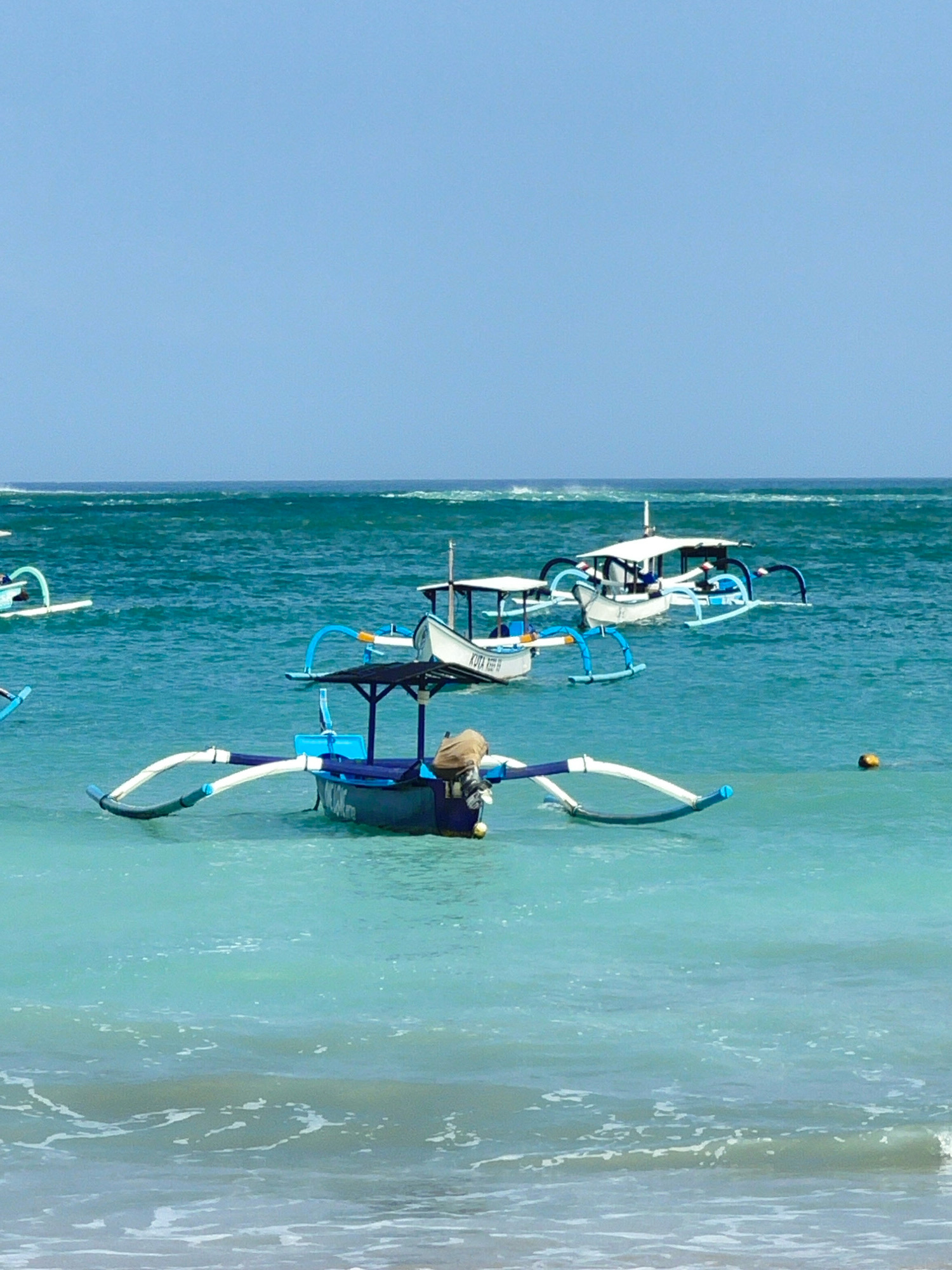 In Kuta, Indonesia, several traditional outrigger boats are moored in the clear turquoise waters of the ocean. The sky is a pale, cloudless blue. The boats, painted in blues and whites, have distinctive outrigger pontoons extending from their sides for stability. One boat in the foreground features a dark blue hull with white lettering and a canvas canopy. A person appears to be seated in this boat, wearing a tan garment and a dark cap. Another boat, further back, is larger and has a white cabin structure with a blue trim. A smaller boat is visible to the left, and another larger boat is in the mid-ground, also featuring the characteristic outrigger design. Gentle waves with white foam are lapping at the foreground, indicating a shallow area near the shore. A small yellow buoy is floating in the water to the right of the foreground boat. The overall scene depicts a tranquil day at sea, with the boats characteristic of coastal Indonesian waters.