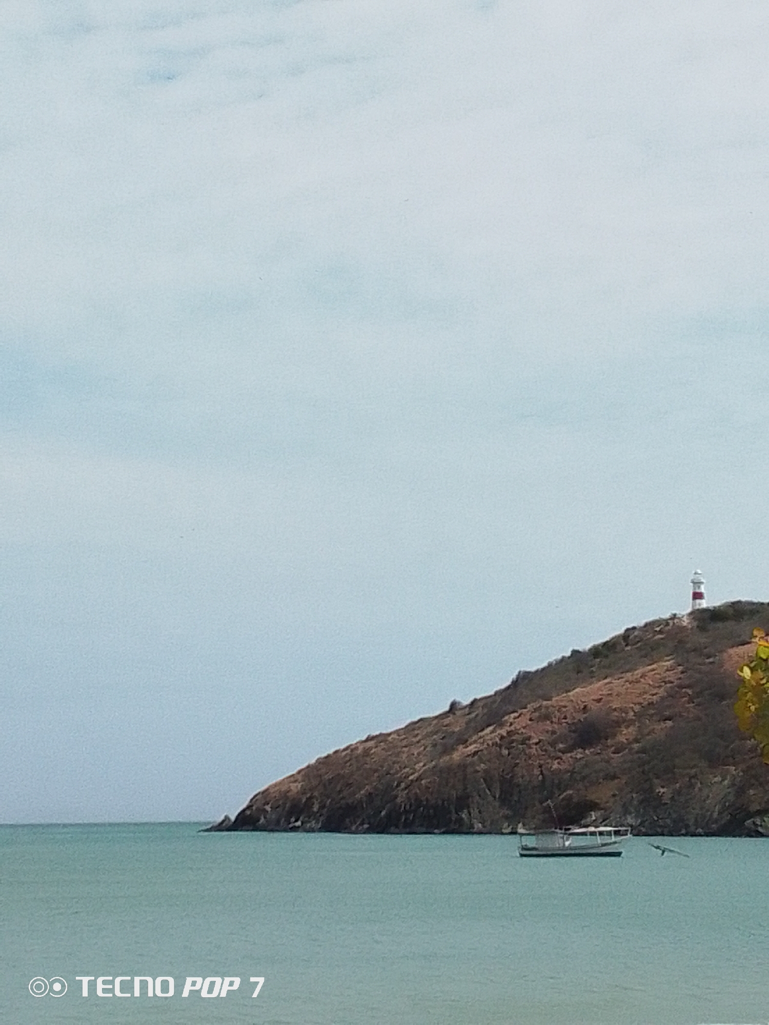 The image depicts a coastal scene in Pedro González, Venezuela. An arid hill slopes into the sea, topped with a small lighthouse painted with red and white stripes. A single small boat is anchored in the water near the hill's base. A bird flies close to the water's surface. The sky is overcast. The image was taken with a TECNO POP 7 phone.
