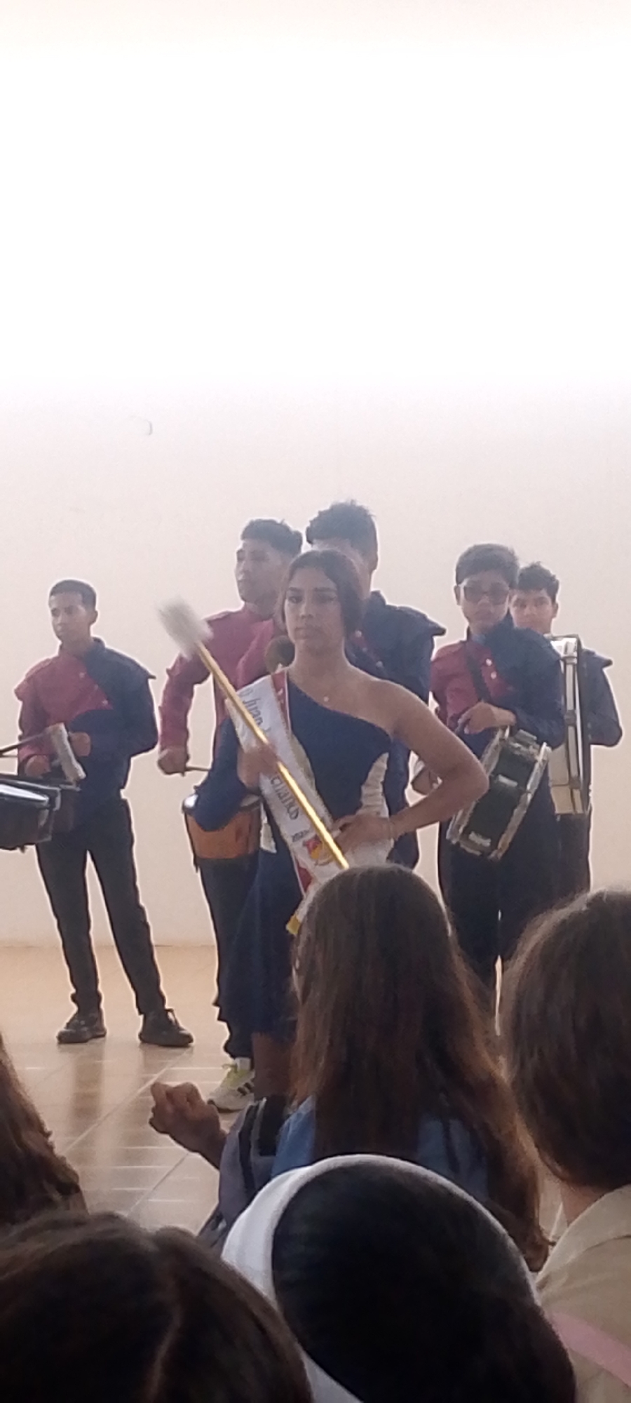 The image shows a group of young people, presumably a school marching band, performing in what appears to be an indoor space, possibly a school gymnasium or auditorium. There are seven people in the marching band. They are wearing uniforms consisting of a red shirt, blue jacket and black pants, and black dress shoes. The band members are playing percussion instruments, including snare drums, bass drums, and other small drums. A young woman is standing in front of the group, wearing a one-shoulder dress. She is holding a baton. It is unclear if this is in Juan Griego, Venezuela. The foreground shows the backs of the heads of audience members.