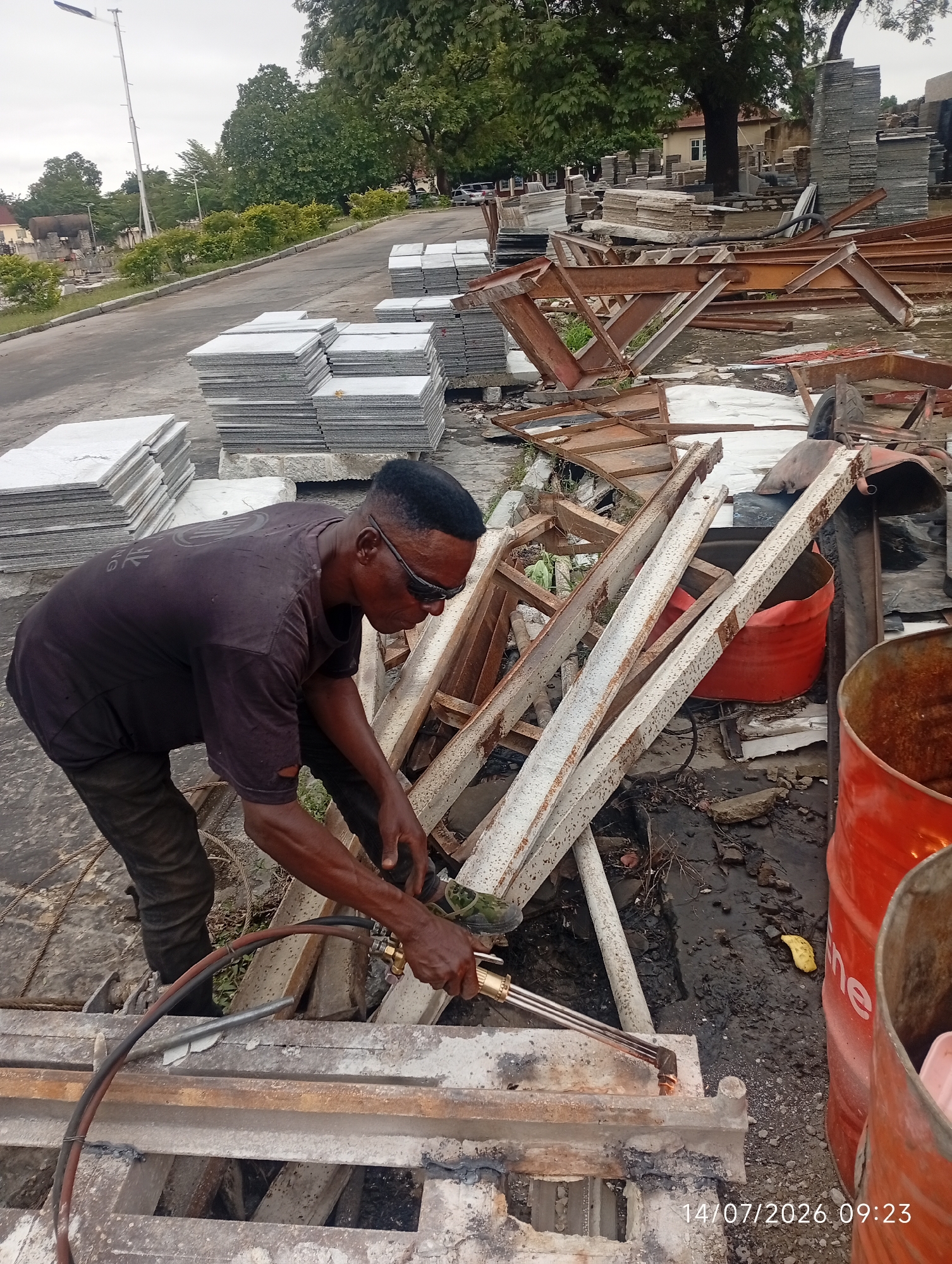 A Black man, wearing a dark t-shirt and dark pants, is seen from the side with his back to the camera, operating a welding torch. He is in the process of welding metal beams, with sparks emanating from the torch. The man is wearing safety glasses. He appears to be in an outdoor area with stacks of what seem to be marble or granite slabs in the background. Several rusty metal structures and large red barrels are also visible. The ground is a mix of dirt and debris. The scene appears to be a workshop or a construction site. A date and time stamp, "14/07/2026 09:23", is visible in the lower right corner of the image. The overall environment suggests a working area with materials and equipment for construction or metal fabrication.