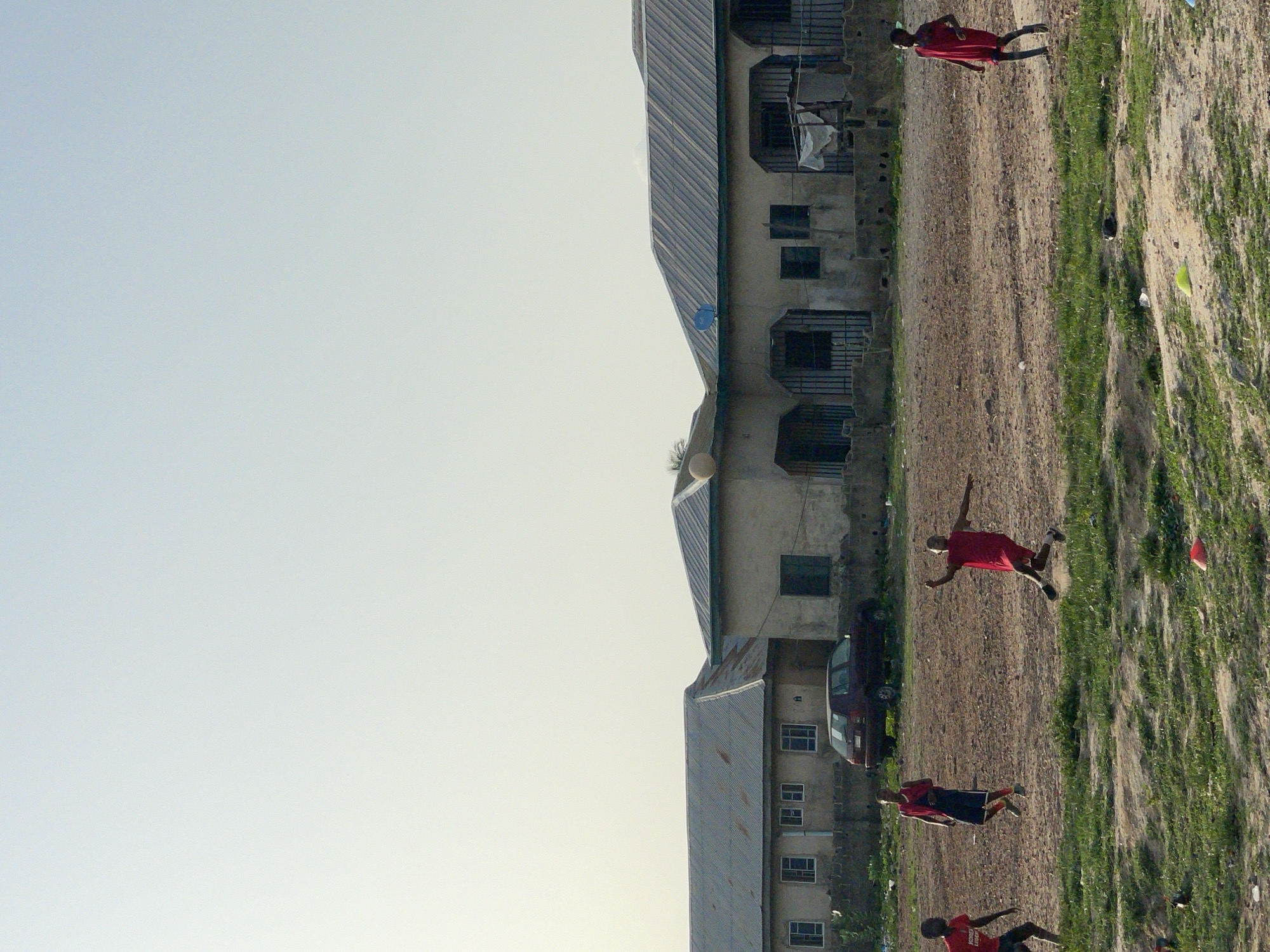 Three people in red clothing jump in a dirt field that is partly covered in green vegetation. They are in front of a multi-story building with a corrugated metal roof and a black car parked nearby. The scene is in Lokoja, Nigeria.