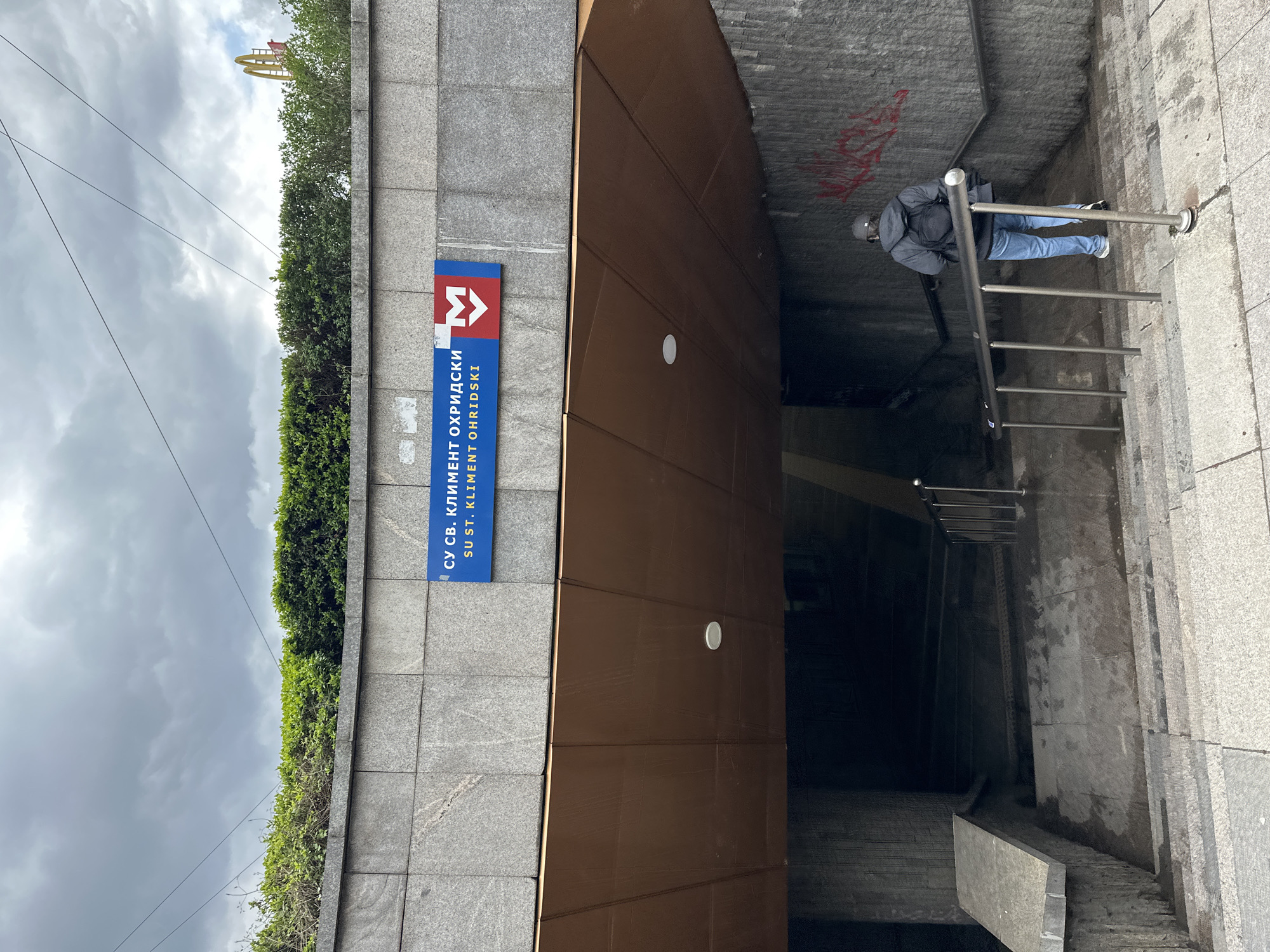 The image shows a ground-level view of a subway station entrance in Sofia, Bulgaria, on an overcast day. The primary focus is the dark, descending stairwell of the metro entrance, flanked by gray, rough-textured stone walls.
A prominent blue sign on the wall to the left of the entrance displays the metro symbol (a red "M" with an arrow) and the station's name. The text is in both Cyrillic and Latin script: "СУ СВ. КЛИМЕНТ ОХРИДСКИ" and "SU ST. KLIMENT OHRIDSKI," identifying it as the St. Kliment Ohridski Sofia University metro station.
One person is visible standing at the top of the stairs, holding onto a silver metal handrail. They are dressed in a dark hooded jacket, blue jeans, and light-colored sneakers, and are carrying a dark backpack. They appear to be in the process of entering or exiting the station. Multiple metal handrails guide commuters down into the dark interior of the entrance. Some red graffiti marks are visible on the ceiling inside the entrance.
Above the entrance, a line of dense green bushes and trees is visible under a heavily overcast sky with gray clouds. In the upper background, partially obscured by the trees, the distinctive yellow "M" arches of a McDonald's sign can be seen, providing a contextual clue for the urban setting. The overall scene depicts a typical city transit point on a cloudy day.