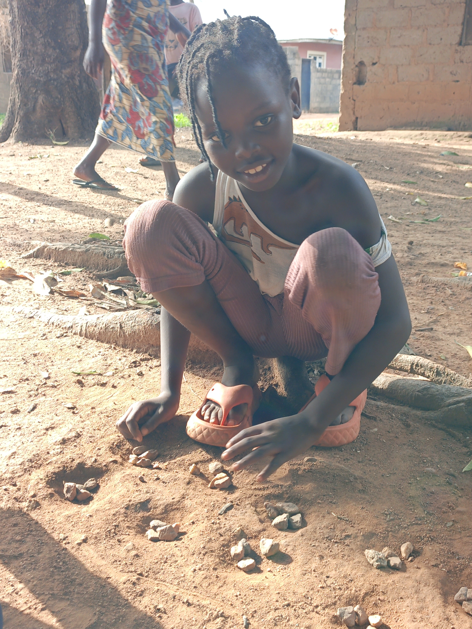 A young African girl is crouched on the ground in what appears to be a rural part of Akwanga, Nigeria. She is wearing a light-colored tank top, reddish-brown leggings, and orange sandals. She's playing a game with small stones, arranging them in groups on the reddish-brown soil. Behind her, a woman in a patterned skirt walks near a tree. In the background, a building with brick walls is visible. The environment is sunny, and the ground is covered in loose soil, stones, and tree roots.