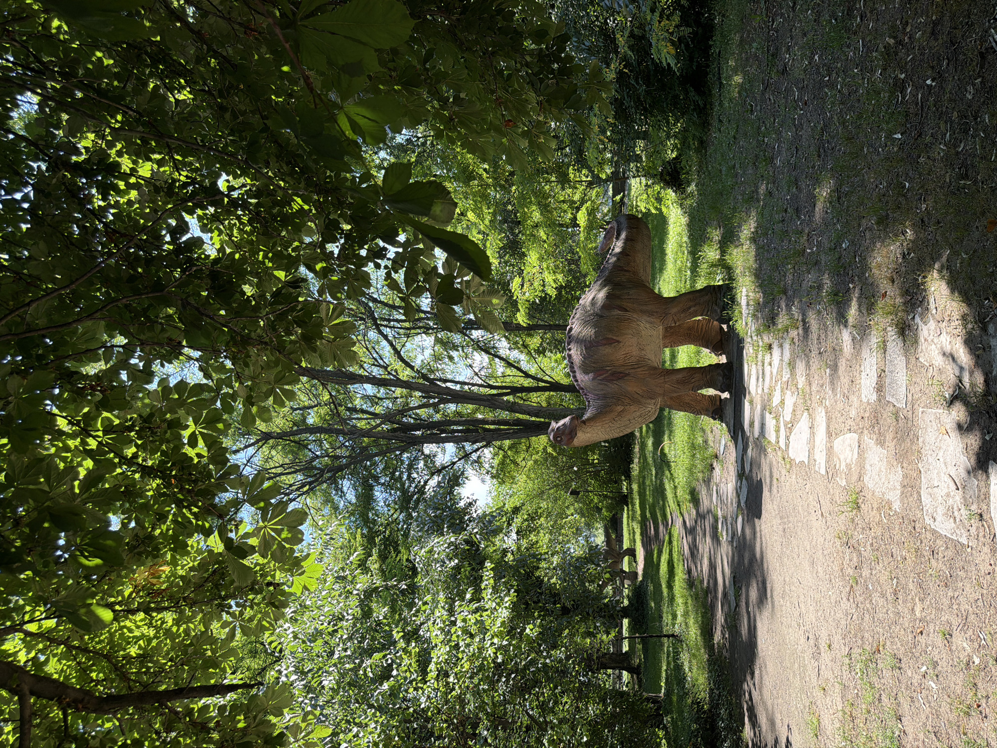 A realistic dinosaur model, resembling a Brontosaurus, stands on a stone path in a park. The dinosaur is light brown with darker markings. The park is lush with green trees and grass, with sunlight filtering through the leaves. A dirt path with stepping stones leads into the background, where another dinosaur model can be partially seen. This scene is located in the Pazardzhik City Park in Pazardzhik, Bulgaria. There are no people or animals visible in the image.