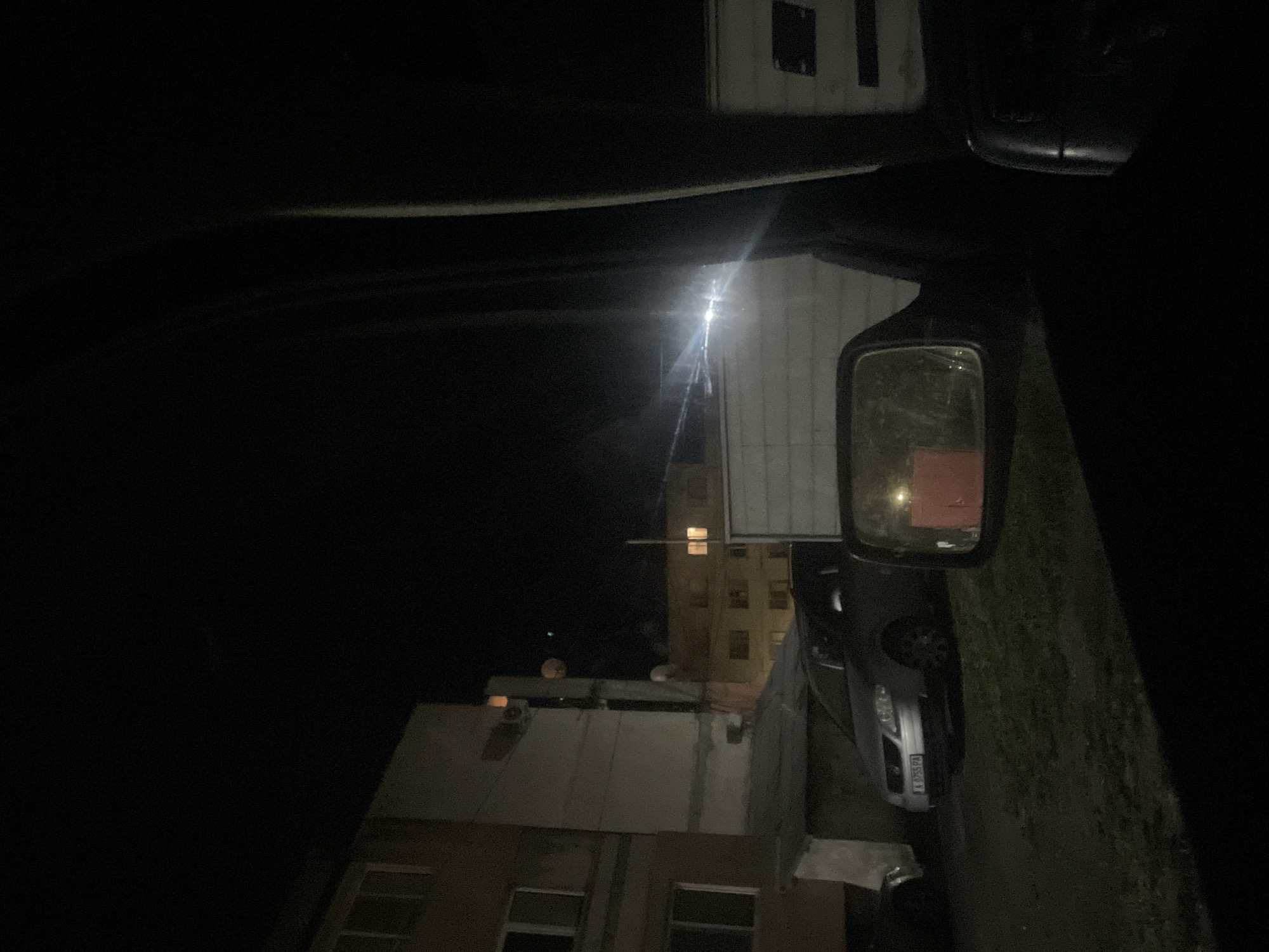 The image shows a nighttime scene in Debelt, Bulgaria, viewed from inside a vehicle, possibly a van. The vehicle's interior is visible along the top of the frame. The view outside includes buildings, a parked car, and a side mirror. The parked car's license plate is partially visible. A streetlight illuminates the area.