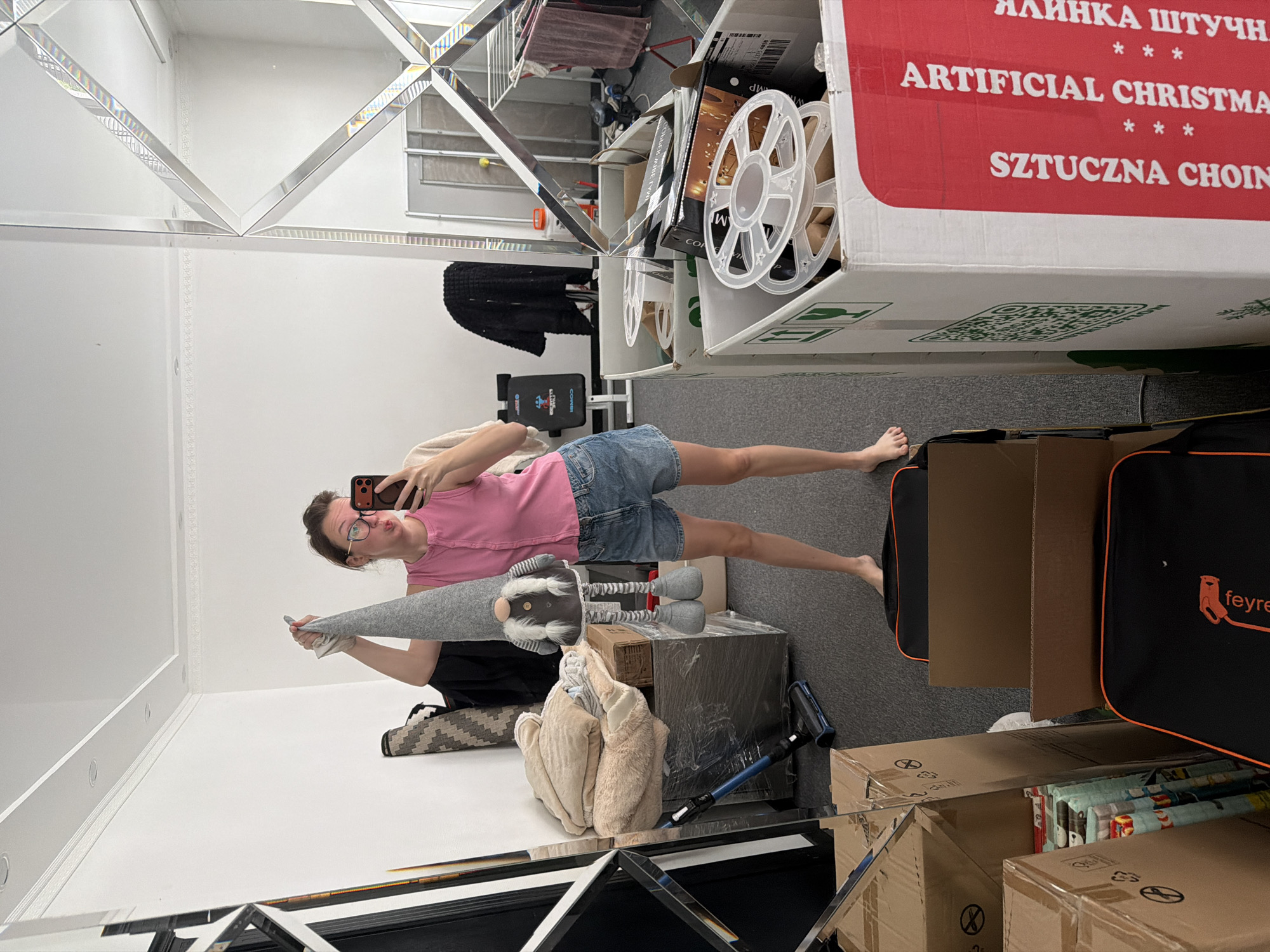 A woman in a pink sleeveless top and denim shorts stands in a room, holding a tall, gray gnome doll by its hat. She is taking a selfie in a large, multifaceted mirror. The room appears to be in a state of disarray, with numerous cardboard boxes, some stacked high, and other items scattered around. To the left of the woman, a vacuum cleaner is visible on the floor. In the background, a weight bench and laundry drying racks can be seen reflected in the mirror. The overall impression is that of a storage area or a room undergoing packing or unpacking. The specific location in Bila Tserkva, Ukraine, cannot be determined from the visual information provided.