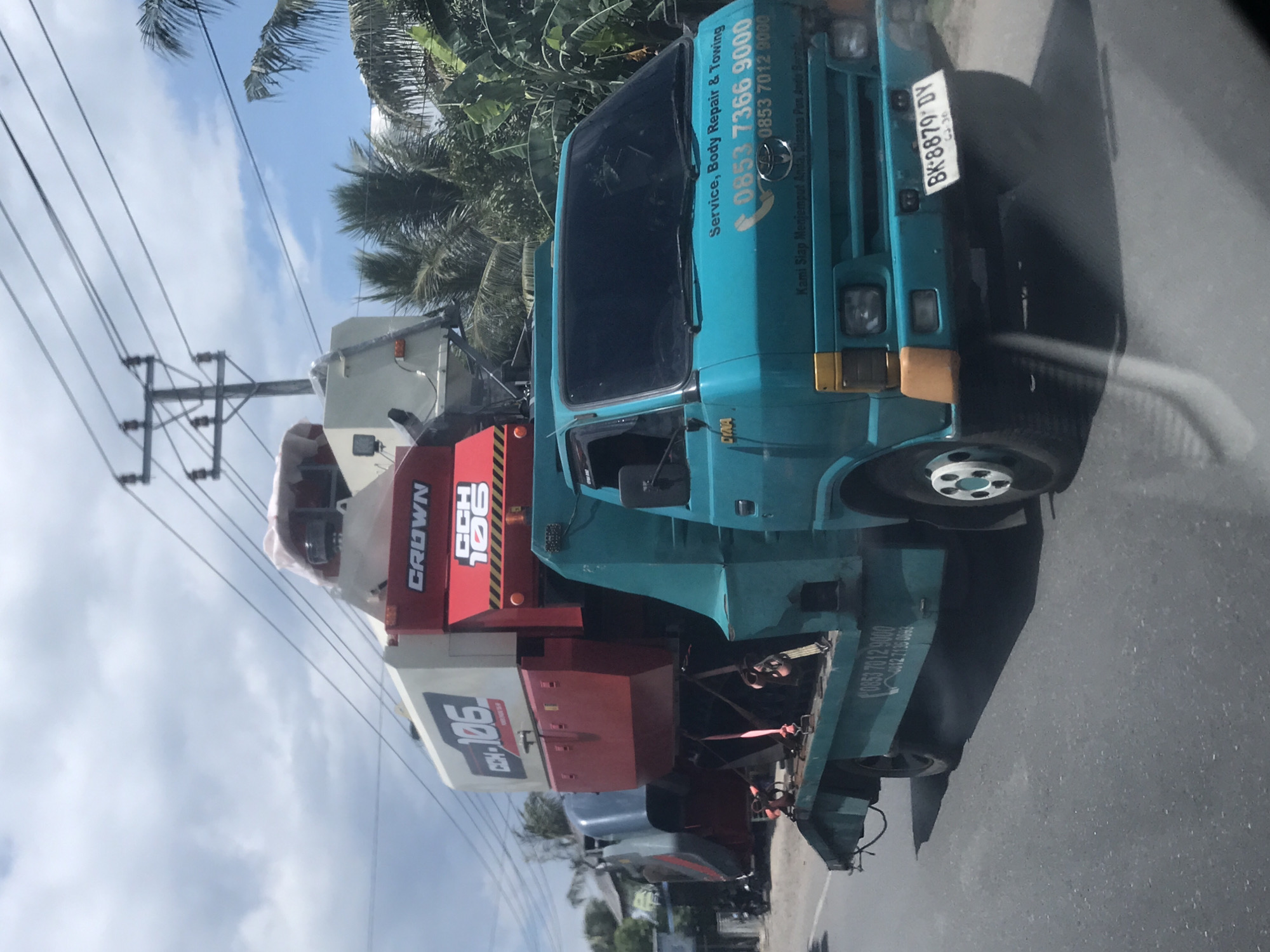 The image depicts a blue Toyota Dyna truck parked on a road, carrying a Crown CCH 106 crane. The truck has a license plate number BK-8879 DY, and signage indicating "Service, Body Repair & Towing" along with a phone number 0853 7366 9000 and 0853 7012 9000. The truck is parked on a road near palm trees and power lines under a partially cloudy sky. Based on the phone number and license plate origin, the image is likely taken in the city of Lhokseumawe, Indonesia.