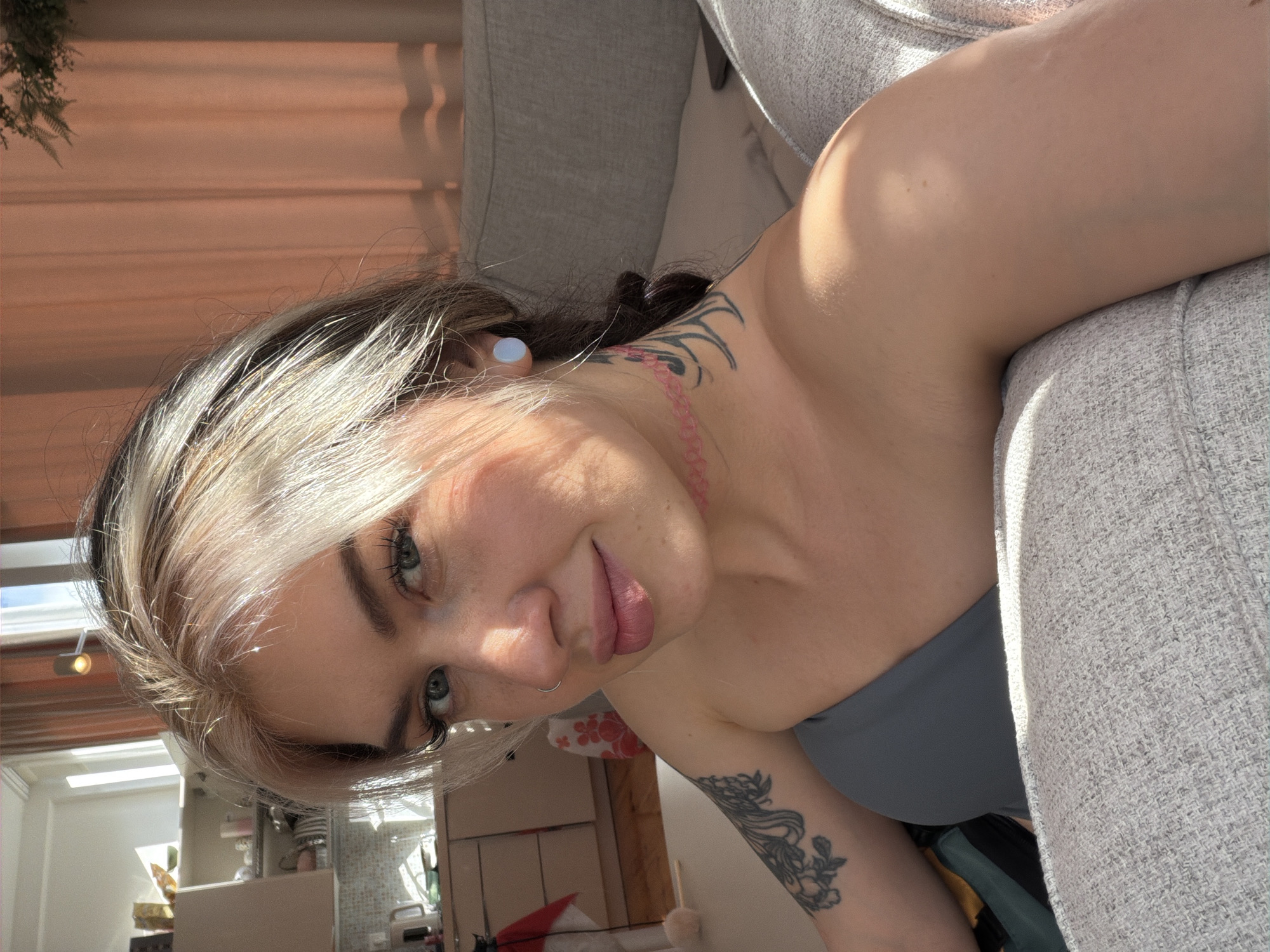 A light-skinned person with blonde and dark hair is lying on a gray fabric couch. The person has blue eyes and a nose piercing. Visible tattoos appear on their neck and upper arm. The setting appears to be inside a residential space, perhaps a home or apartment, based on the presence of a kitchen area in the background. The image does not provide enough information to determine a specific location in Budva, Montenegro.