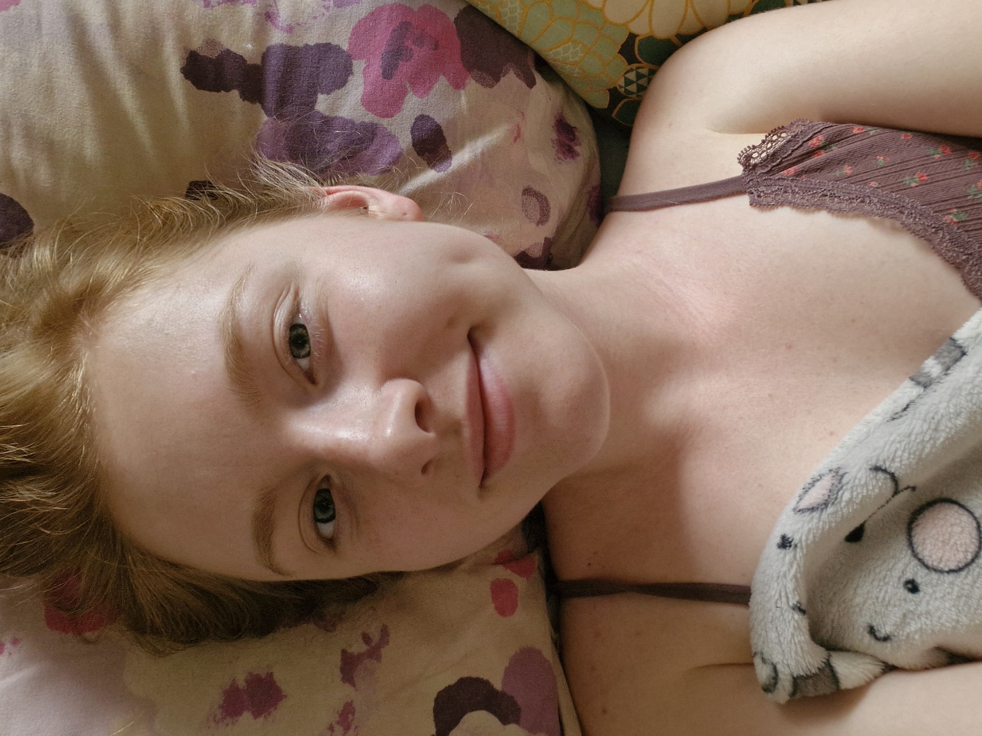 A fair-skinned young woman with reddish-blonde hair lies on a pillow patterned with purple and pink floral designs, partially covered by a fuzzy gray blanket with a cartoon animal face. She is wearing a brown camisole with lace trim. The location is not discernible from the image.