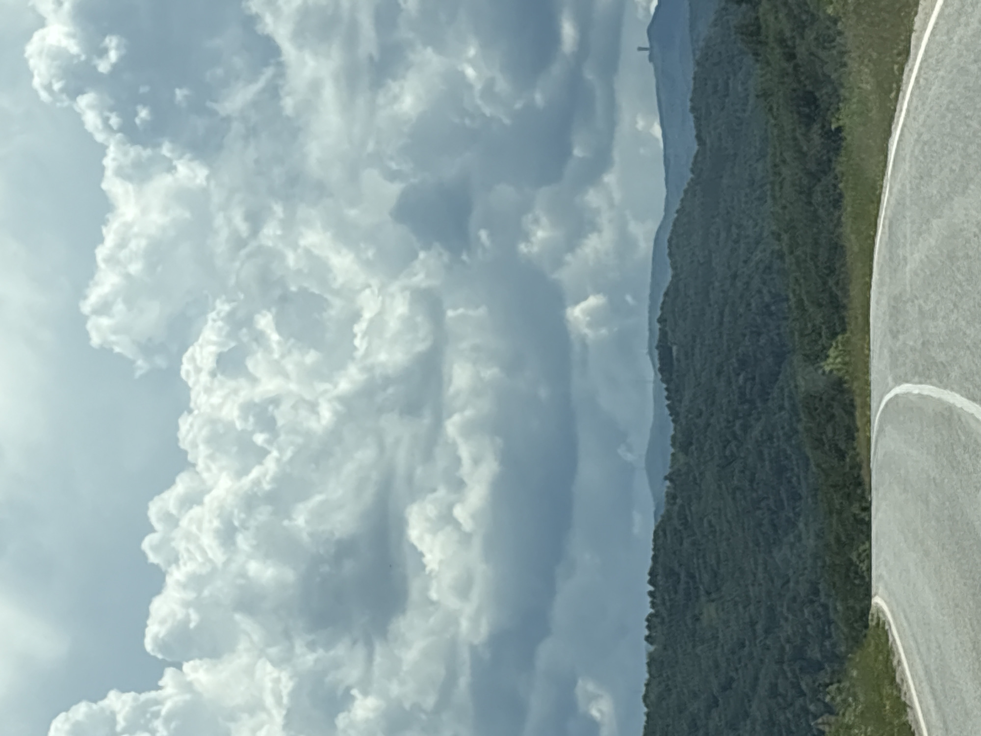 The image depicts a winding asphalt road in the foreground, with white lane markings curving to the right. Beyond the road, rolling hills covered in dense green forest stretch into the distance. In the far background, silhouetted against a cloudy sky, are a series of mountain peaks, one of which features a tall, slender structure, possibly a tower or communication antenna. The sky is filled with dramatic, voluminous clouds, with patches of brighter sky visible above. The overall impression is of a rural, mountainous landscape under a dynamic sky. The scene is consistent with the views from roads leading out of the city of Gabrovo, Bulgaria, towards the Stara Planina mountains, where such towers are often situated on prominent peaks.
