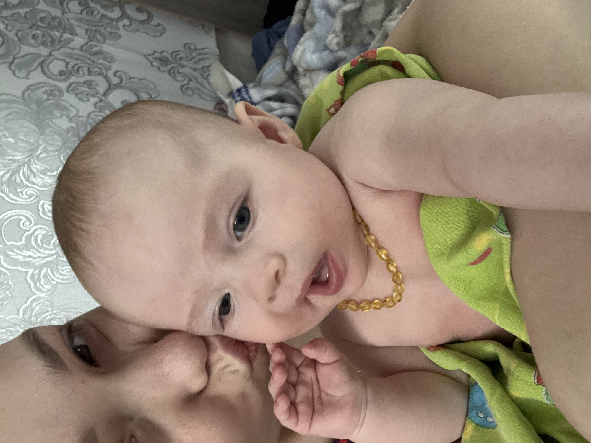 A baby with an amber teething necklace lies on a bed with patterned bedding, partially wrapped in a green patterned blanket. A woman's face is close to the baby, appearing to kiss the baby's cheek. The location is Кемуль, Russia.