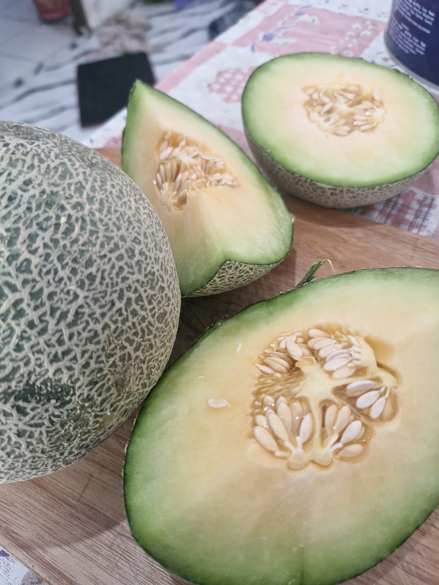 The image shows a whole cantaloupe and several slices of cantaloupe arranged on a wooden cutting board. The cantaloupe has a textured rind with a pattern of light and dark green lines. The flesh of the melon is a light orange color, and the seeds are visible in the center of the slices. The background is blurred but appears to be a patterned surface, possibly a tablecloth. There is no indication of the city of Rantau, Indonesia, in the image or its context.