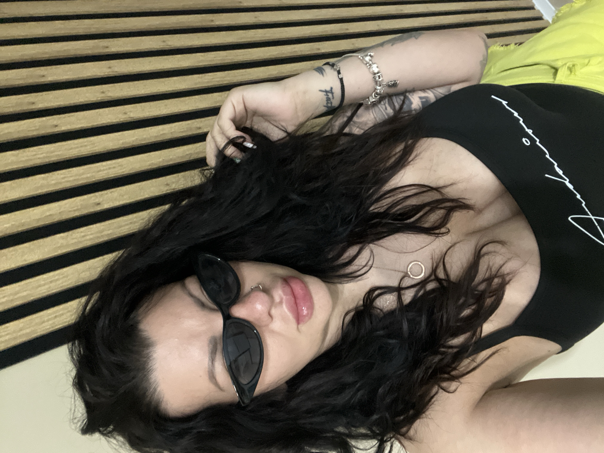 A woman with dark, wavy hair is reclining, wearing sunglasses, a black tank top, and yellow shorts. She has multiple tattoos and bracelets on her left arm, and a circular pendant necklace. The background features a wall with alternating dark and light brown horizontal stripes. It is not possible to infer the location of the photo, including whether it's in Plovdiv, Bulgaria, with the given information.