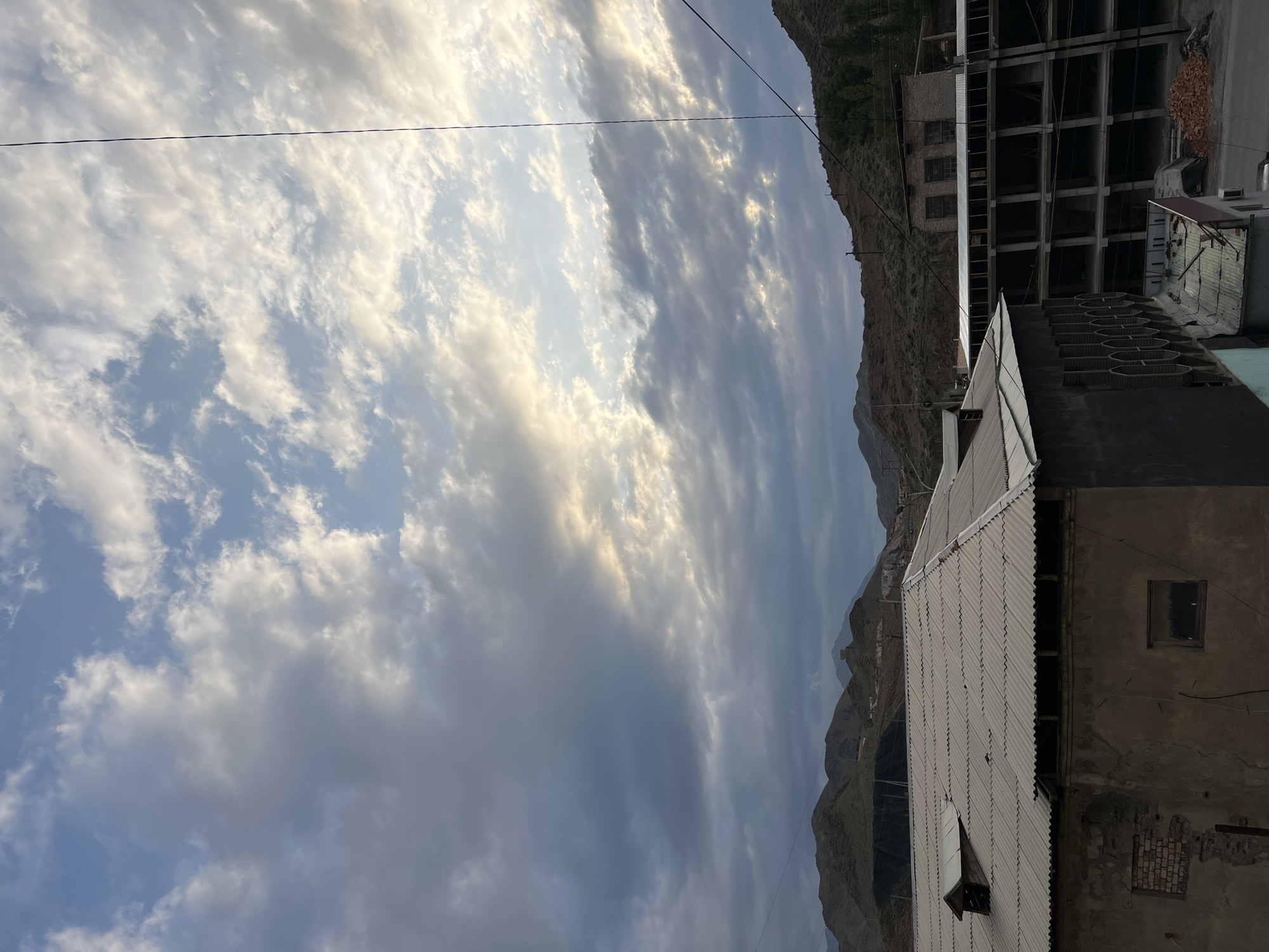 The image depicts a cloudy sky over a somewhat rural or industrial area in Kadamjay, Kyrgyzstan. In the foreground, there are buildings with corrugated metal roofs. One building is older, with a worn facade and exposed brick in places, and appears to have a small dormer window on its roof. Adjacent to it is a multi-story structure that appears to be under construction, with concrete floors and supporting columns visible, and a pile of bricks on the ground. The background features rolling hills and mountains, characteristic of the landscape around Kadamjay. The lighting suggests it is either early morning or late afternoon, with the sun breaking through the clouds in some areas. There are no people visible in the image. The scene is quiet and appears to be a moment of observation of the environment.