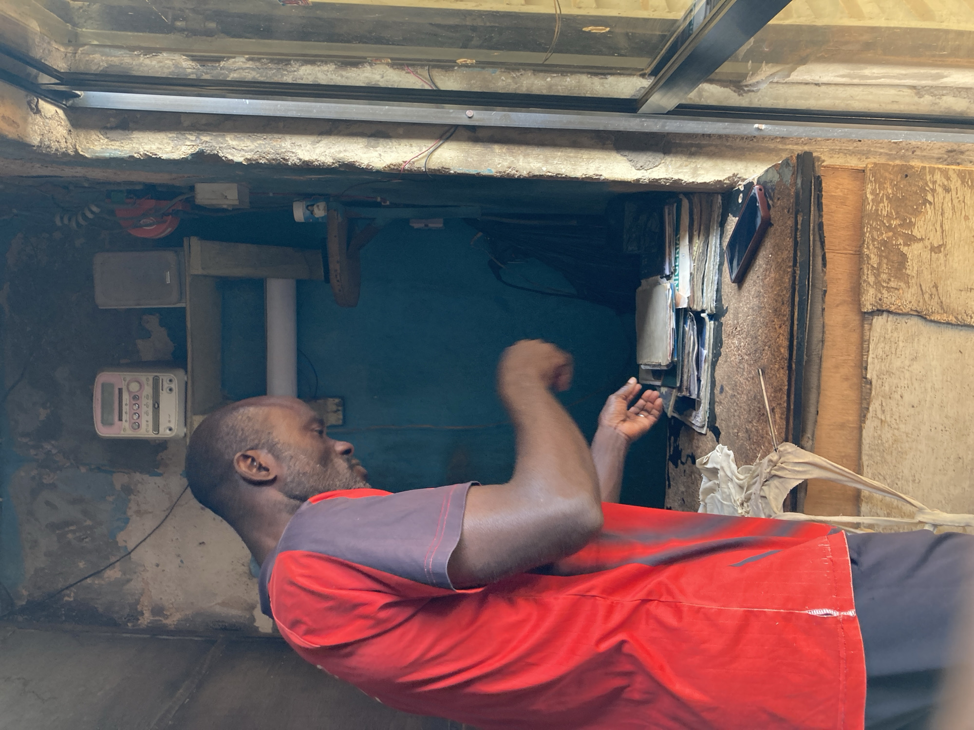 An image depicts a man in Jos, Nigeria, in what appears to be an indoor utility area. The man is seen from the side, wearing a red and gray shirt. His arms are raised. To his left is a wall with a utility meter and other devices. Overhead is the ceiling with metal support structures. The walls appear to be concrete. To the right is a partial wall constructed of wood. Cables and wires are seen throughout the space.
