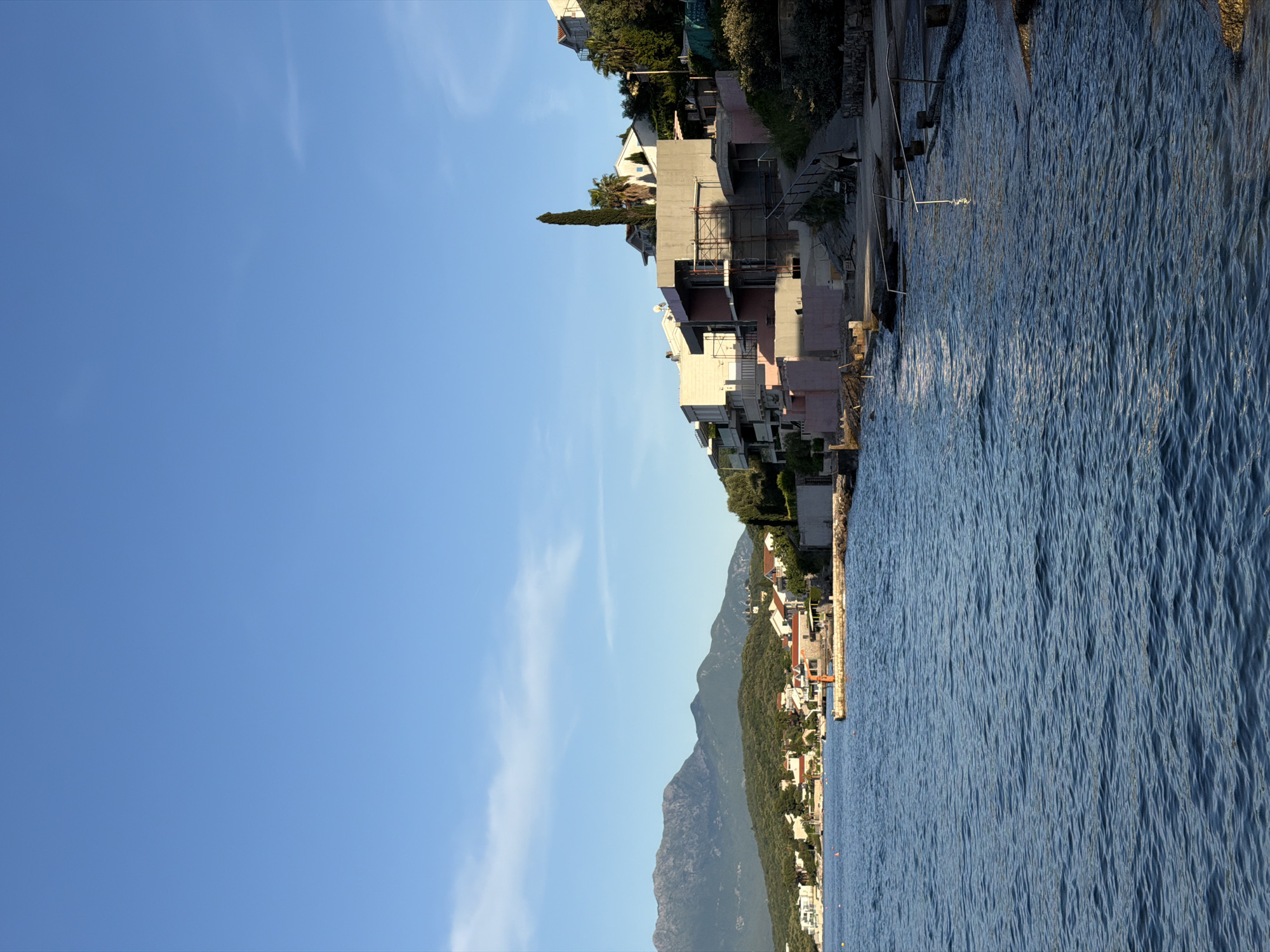 The image shows a coastal scene in Krašići, Montenegro. The view is divided between a clear blue sky on the left and the Adriatic Sea on the right. A line of buildings and mountains runs along the horizon, separating the sky and sea. Closer to the top, construction is visible near the water.
