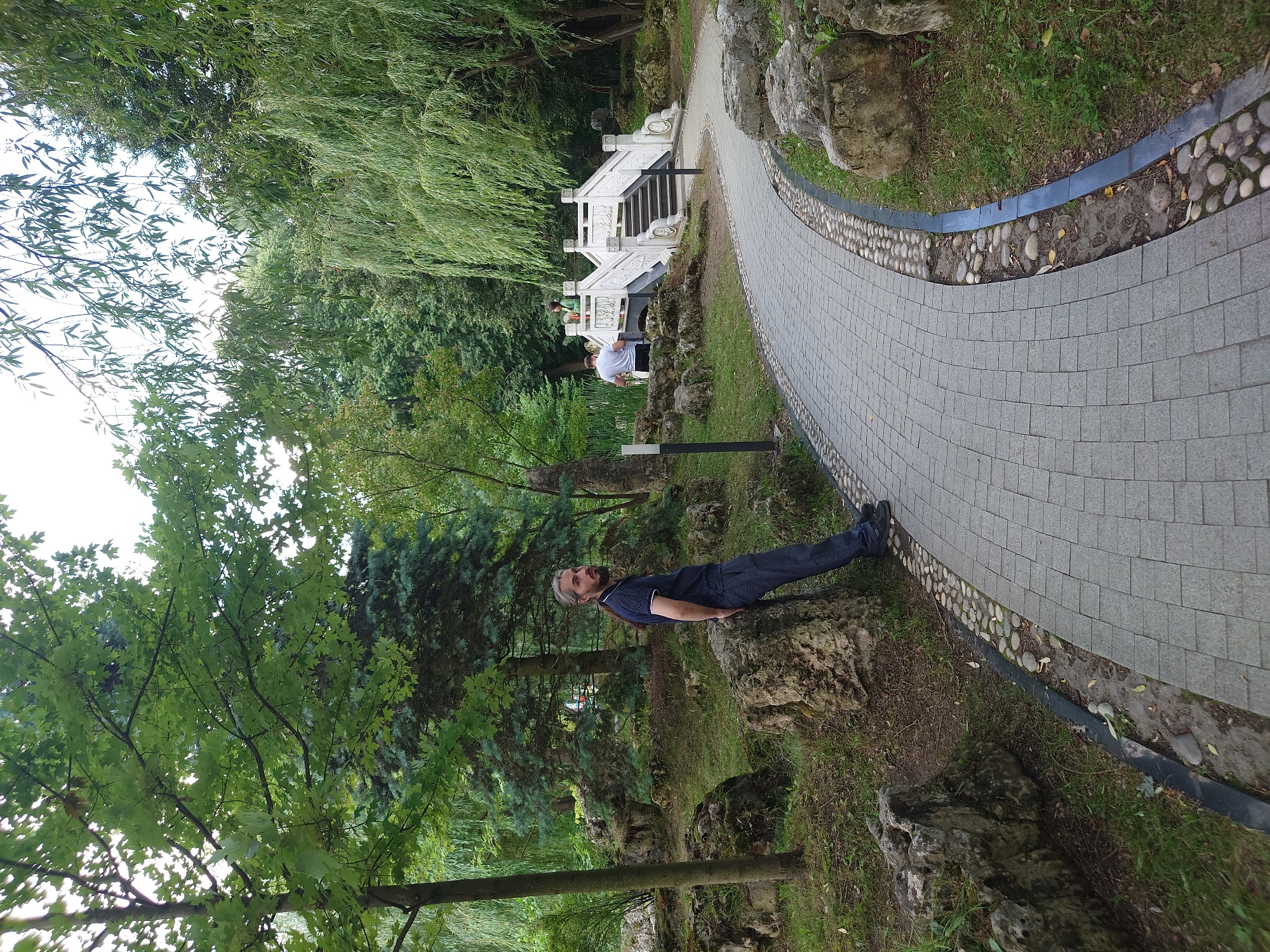 A man with long, graying hair is leaning against a large rock in a park. He is wearing a dark blue polo shirt with white trim on the collar and sleeves, and dark gray pants. The park features a winding pathway paved with grey square tiles, bordered by small, rounded pebbles. To the right of the pathway, a lush green lawn is dotted with rocks and trees. In the background, a white stone bridge with intricate carvings arches over a body of water. Two individuals, one in a white shirt and another in a green jacket, stand on the bridge. The environment is verdant, with mature trees forming a canopy overhead. The scene appears to be located in a botanical garden or a park designed with Asian architectural influences. This image was taken in the Moscow State Botanical Garden of the Russian Academy of Sciences, Moscow, Russia.