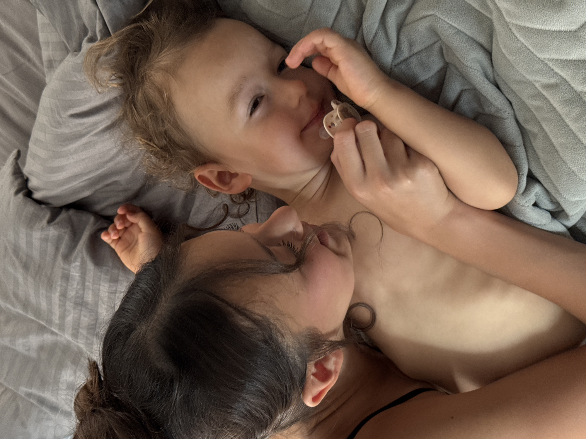 This is an overhead shot of a mother and her young child lying in bed. The mother, with dark hair and an ear visible, is nestled close to the child. The child, with light brown curly hair, is lying on their back and has a pacifier in their mouth, held by their hand. Their eyes are partially closed and they appear content. They are both lying on a grey striped duvet. The lighting suggests it might be daytime, but the exact time is unclear. The location context is Sochi, Russia.