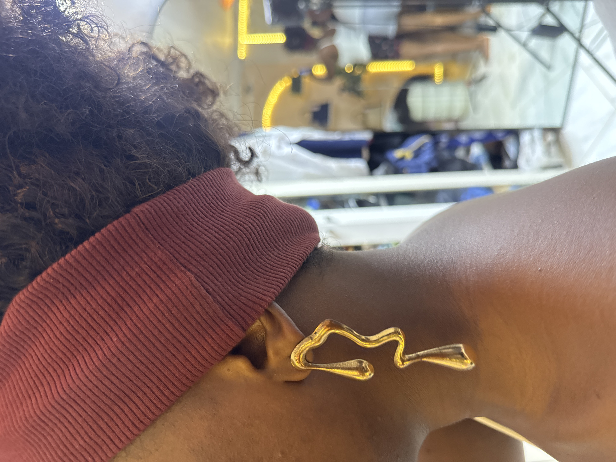 A close-up profile shot shows a person wearing a maroon ribbed headband and a large, gold, abstract-shaped earring in their left ear. The background is blurred but appears to be an interior space with a mirror reflecting yellow lighting and the lower half of another person. The scene is likely indoors, possibly in a retail environment or a personal dressing area. No specific location in Abuja, Nigeria can be determined from the visual information.