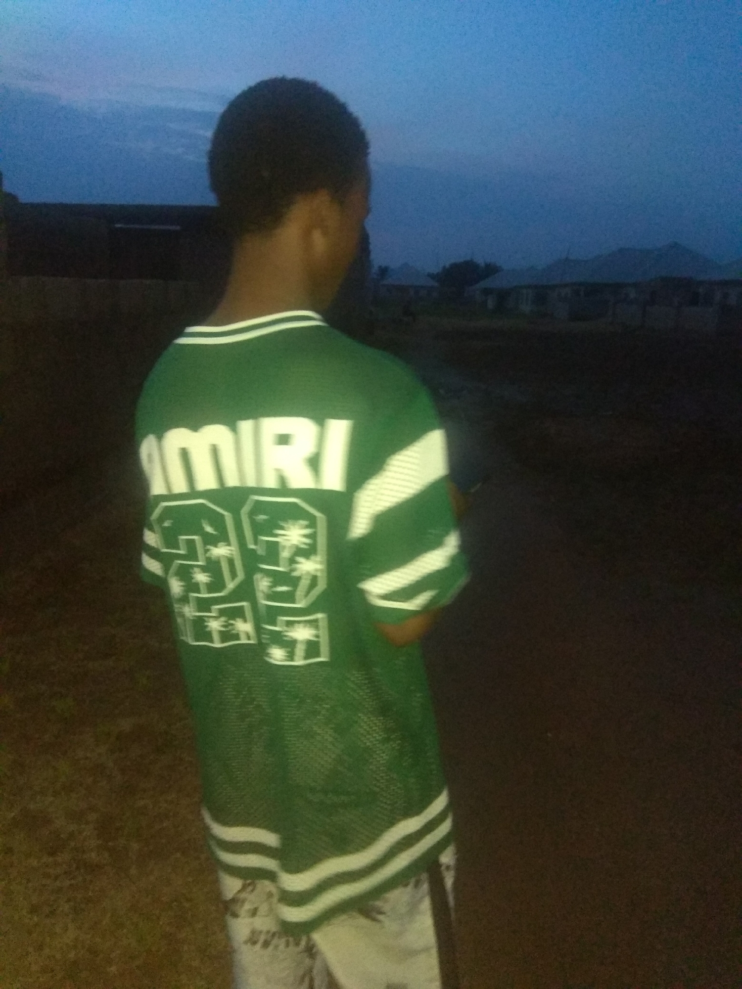 A person, seen from the back, stands on a dirt road at dusk in a residential area of Jalingo, Nigeria. The person is wearing a green mesh jersey with white lettering that spells "AMIRI" across the back. Below the name are two large white numbers, "23", each containing a graphic of palm trees. The jersey has white stripes on the sleeves and collar. The person is also wearing light-colored shorts. In the background, several houses with dark roofs are visible, along with scattered trees. The sky is a deep blue, indicating the time of day is early evening. The overall scene is dimly lit, with the focus on the person and their attire.