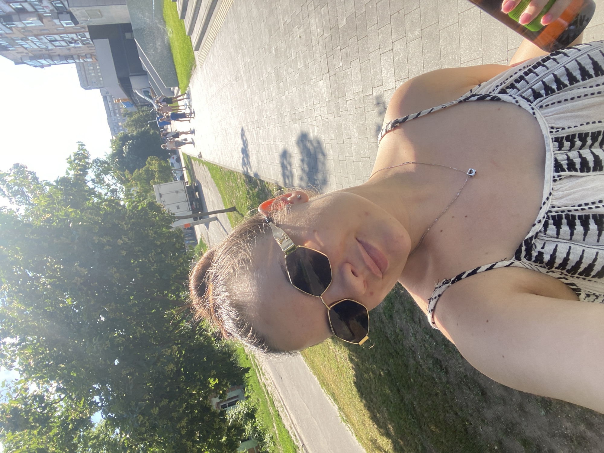 A woman is taking a selfie outdoors during the daytime. She is wearing sunglasses and a patterned dress. In her right hand, she holds a bottle. The background shows a paved walkway, green grass, trees, and buildings. There is a group of people walking in the distance along the walkway. The scene appears to be a city park or public space on a sunny day.