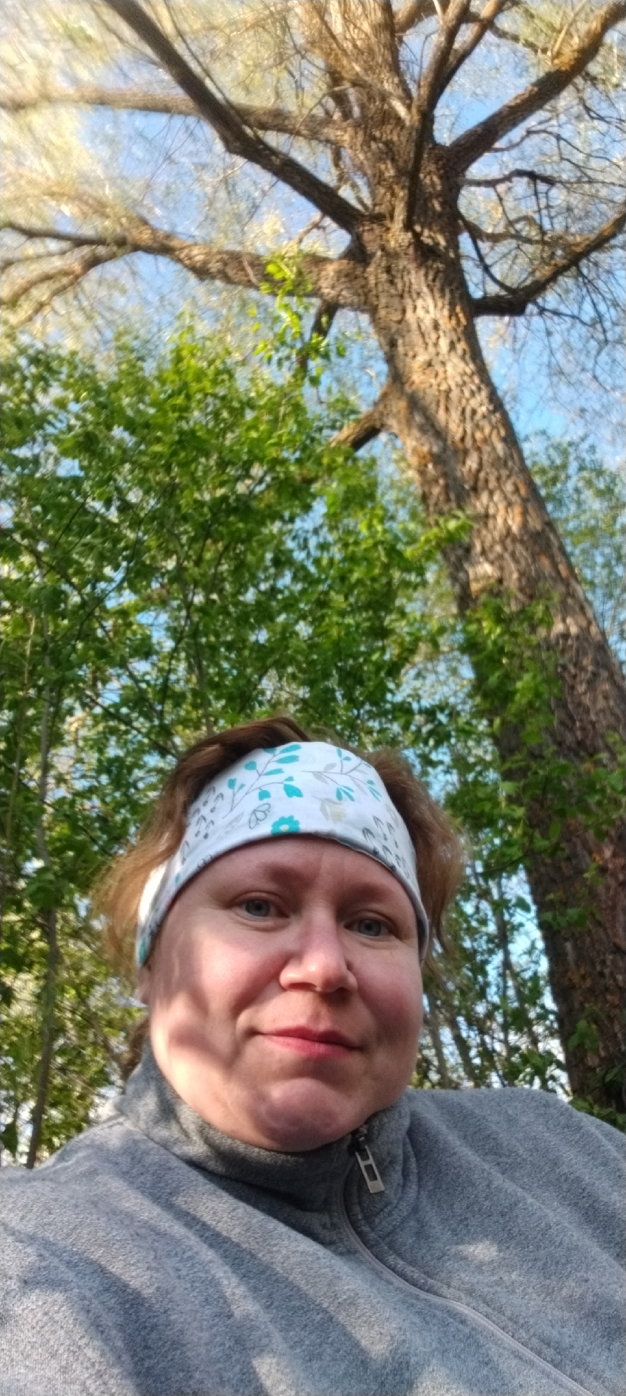 A woman wearing a gray zip-up jacket and a patterned headband is taking a selfie outdoors. Tall trees surround her, some with green leaves and others with bare branches against a blue sky. The location is Зипуново, Russia.