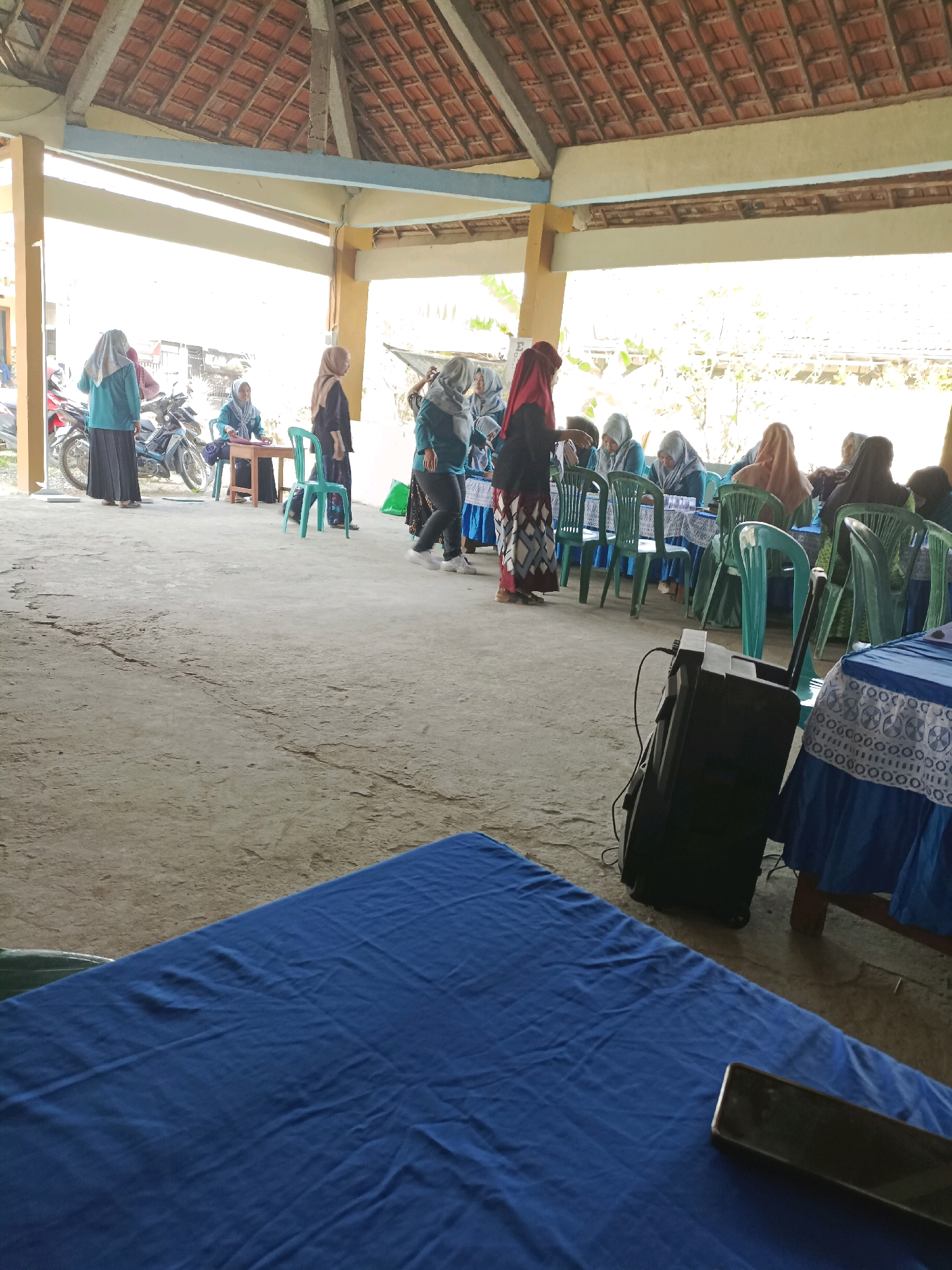 The image shows an outdoor gathering or event at what appears to be a community hall or pavilion in Rancang Kencono, Indonesia. In the foreground, a blue tablecloth covers a table. Behind this, a black speaker is positioned on the ground. Further back, several people, predominantly women wearing hijabs and long skirts or dresses, are seated at tables covered with blue tablecloths. Some are engaged in conversation, while others appear to be listening or observing. A few individuals are standing, including one woman wearing a teal top and black skirt and another in a red hijab and black attire. Motorcycles are visible in the background on the left side of the image. The environment is partially covered by a tiled roof supported by pillars, with the sky and vegetation visible in the distance. The ground surface is concrete.
