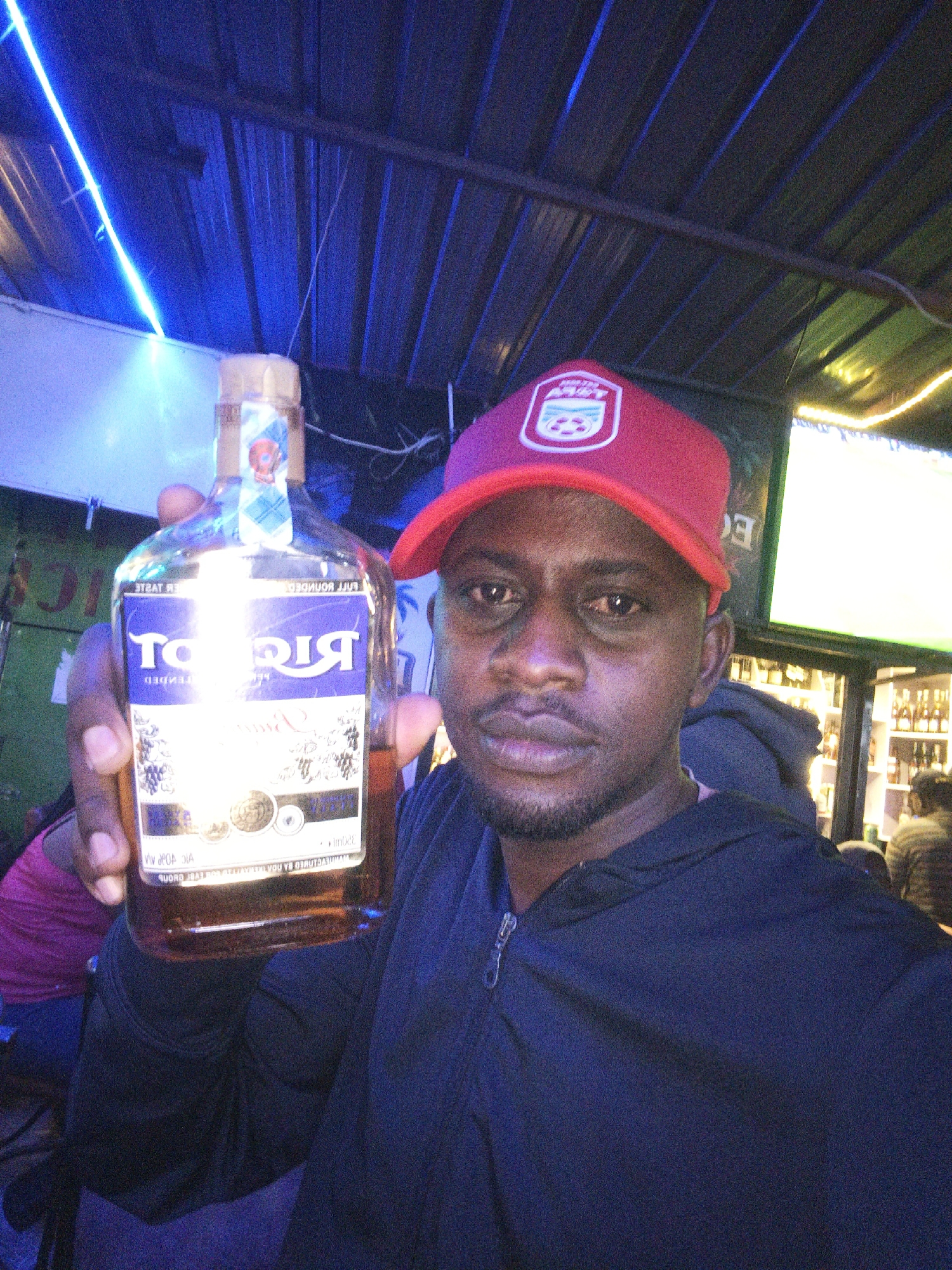 A man wearing a red cap and a navy-blue zip-up hoodie is holding a bottle of Ricot brandy. The brandy label is mostly purple and gold. He is inside what appears to be a bar or lounge. The lighting is blue, and there are lights on the ceiling. Shelves of alcohol bottles are visible in the background, as well as patrons. Based on the appearance of the bottles, style of clothing, and architectural elements like corrugated roofing and lighting, this image might be in a bar or lounge located in Nairobi, Kenya.