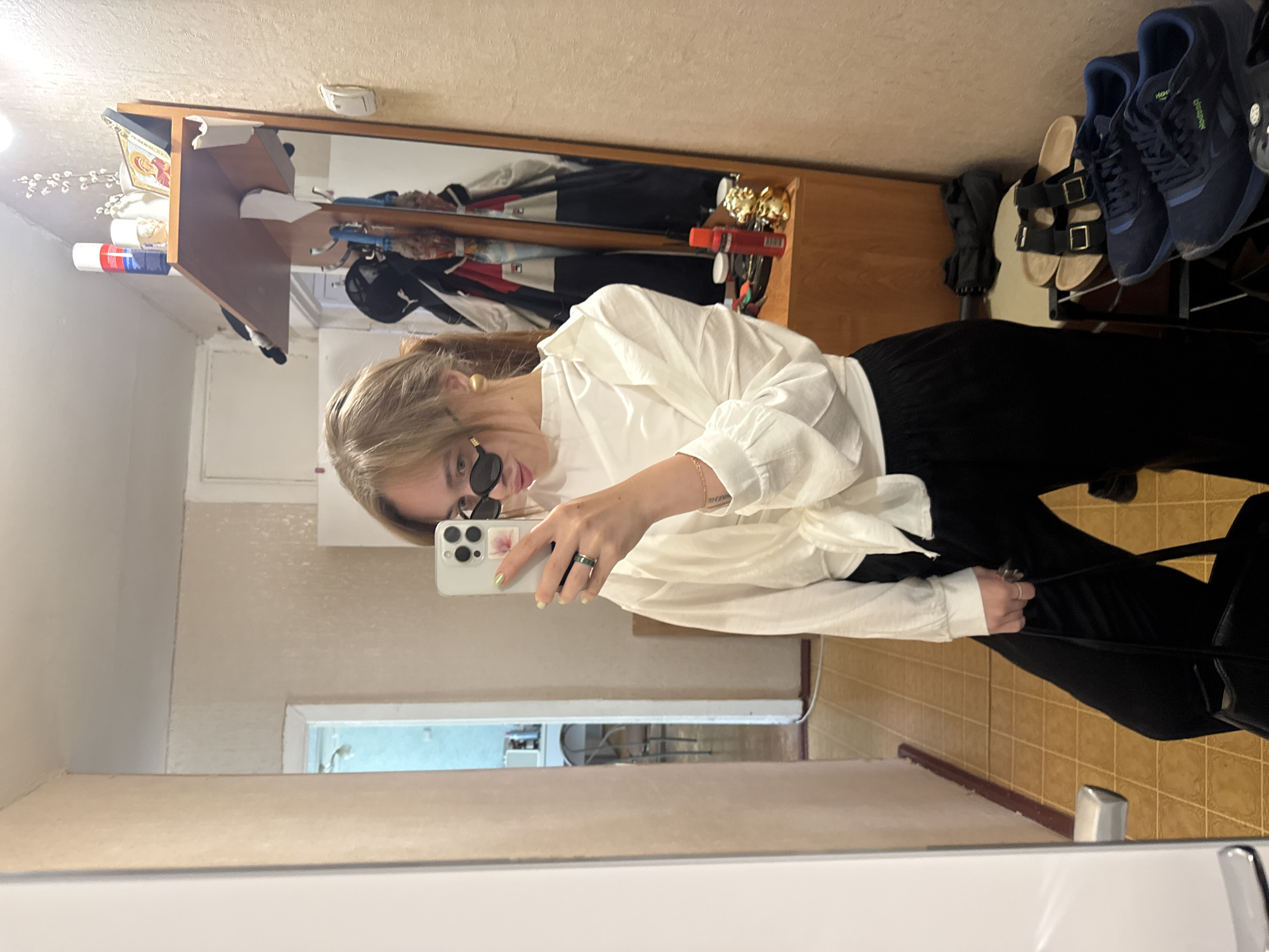 A young woman is taking a selfie in front of a mirror. She is wearing sunglasses, a white shirt, and black pants. The reflection in the mirror shows a hallway with a door at the end and various objects placed on a wooden shelf including, shoes, clothes, and decorative items. The location is Chaikovsky, Russia, judging by the subject's tattoo reading "Чайковский". The floor is tiled. The woman is holding a smartphone.
