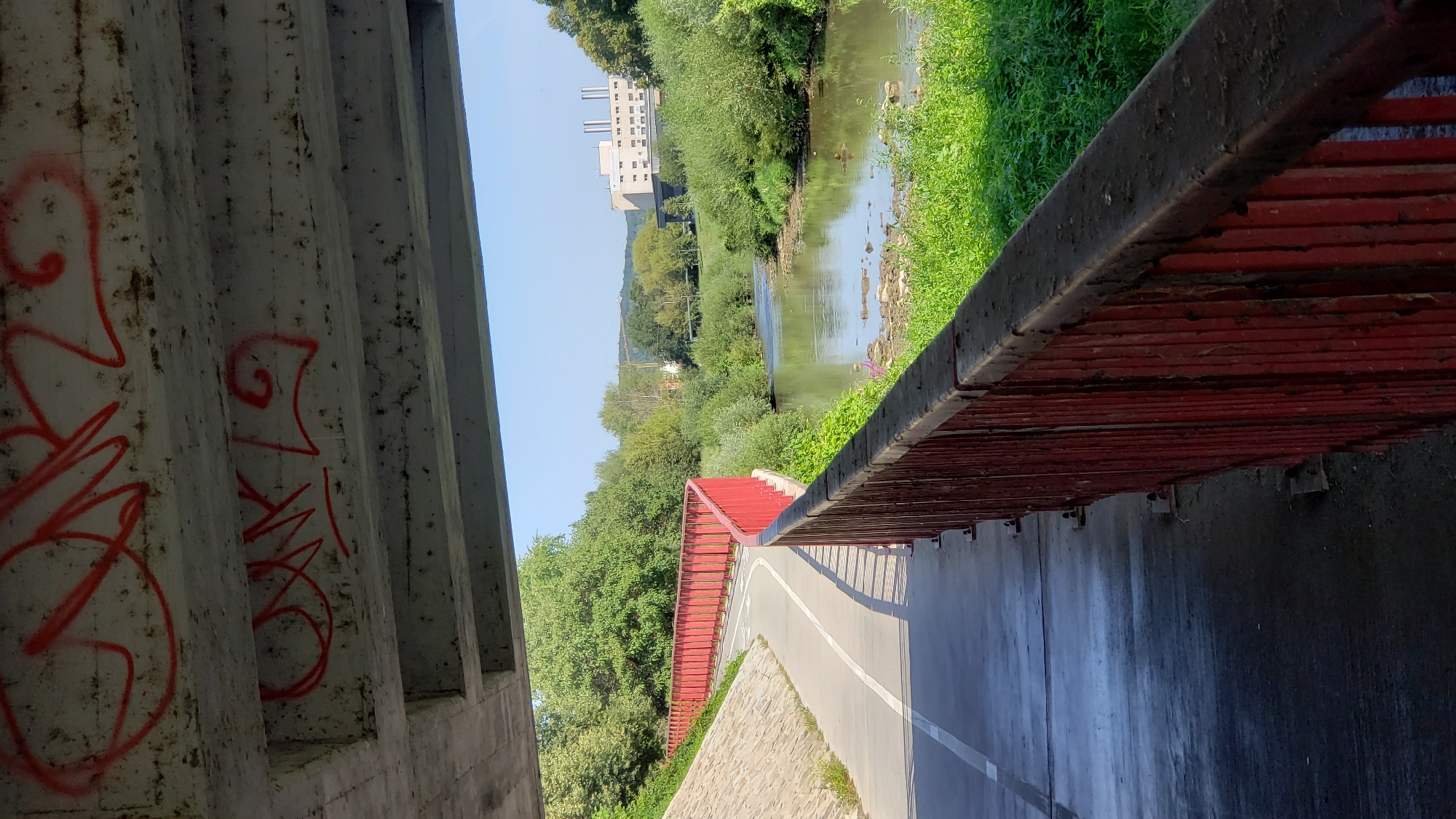 Underneath a concrete overpass, a paved pathway curves to the left, bordered by a red metal railing. The pathway has a white line marking a bike lane. To the right of the pathway and railing, a river flows. The riverbanks are overgrown with green vegetation. In the distance, across the river, are trees and a light-colored industrial building with multiple smokestacks. Red graffiti is visible on the underside of the concrete overpass. The scene appears to be outdoors during the daytime, with a clear blue sky. The location is the cycling path along the Torysa River in Prešov, Slovakia, specifically under the bridge that carries the road from Sekčov towards the city center.
