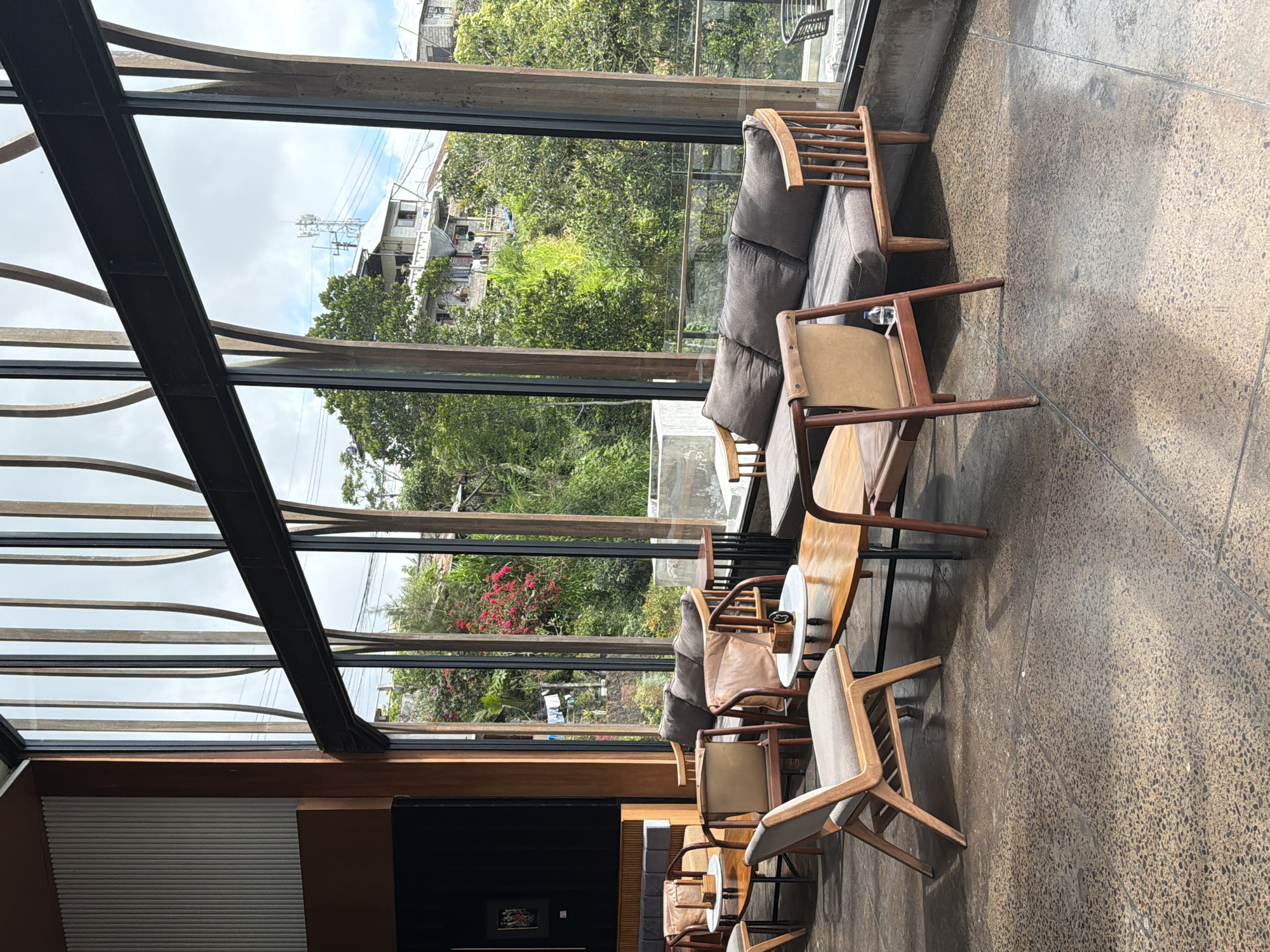 The image depicts the interior of a cafe with large windows offering a view of a lush, hilly landscape in Kintamani, Indonesia. The cafe features modern, minimalist furniture, including several wooden chairs with upholstered seats and a grey fabric sofa. Coffee tables with wooden tops and dark metal legs are placed in front of the seating arrangements. The floor is a polished concrete with aggregate. The windows are framed by dark metal beams and vertical wooden elements that create a distinct architectural style. Outside, a dense green hillside is visible, with some buildings and a telecommunications tower perched on top. Pink flowers are also scattered among the greenery. The scene is well-lit by natural light filtering through the windows. No people are visible in the image.