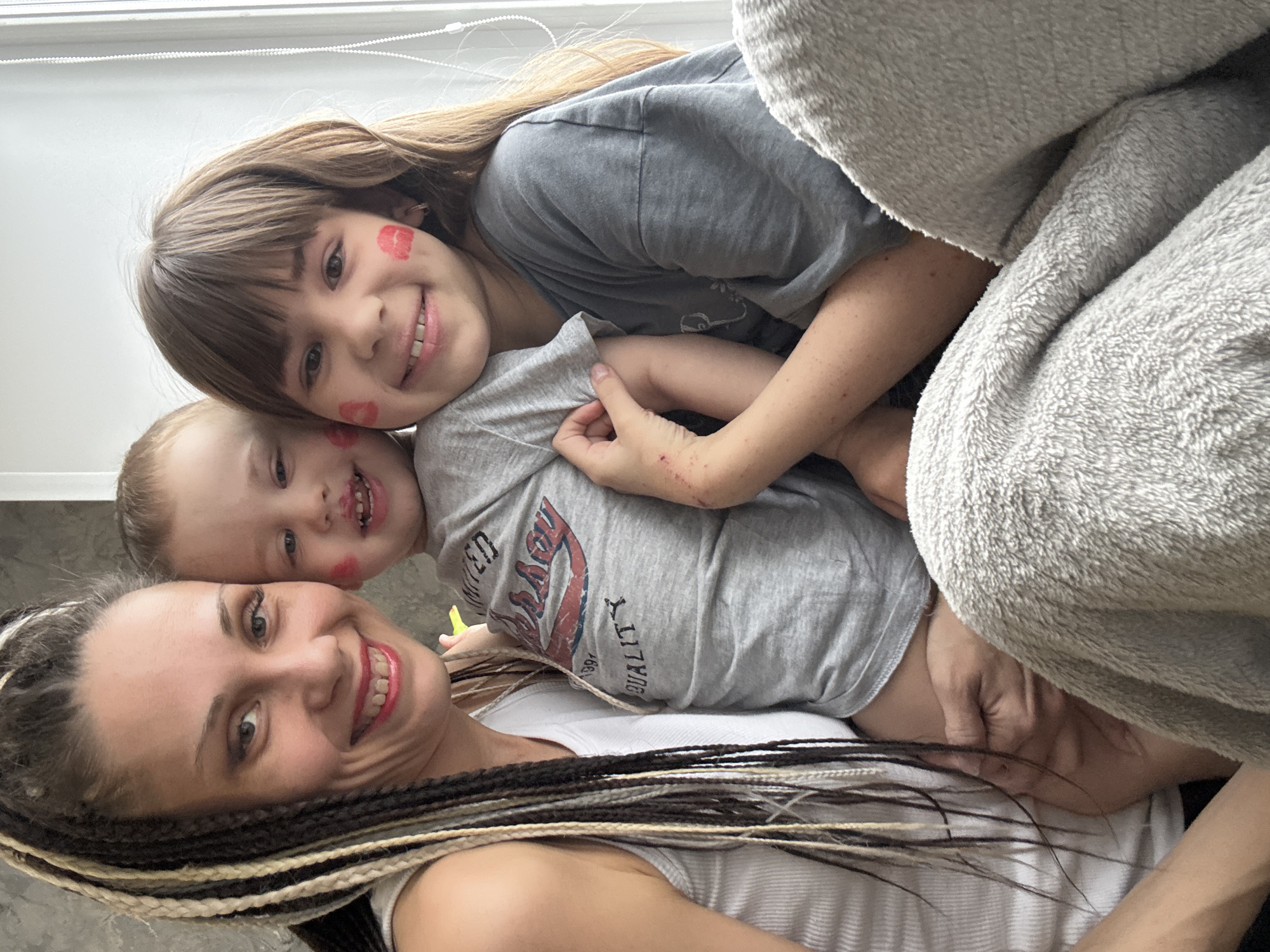 This close-up portrait-oriented photograph, taken indoors, features an adult woman and two children—a young girl and a toddler boy—all smiling joyfully at the camera. The setting suggests a cozy, personal space, possibly in Rusanovo, Russia.
At the bottom of the frame, the adult woman smiles broadly, showcasing a happy expression. She has distinct dark and blonde braided hair. Above her, a young girl with long brown hair and bangs smiles, her cheeks adorned with playful red kiss-mark imprints. She is wearing a grey t-shirt. The toddler boy, nestled between the two, also grins widely, with similar red kiss marks on his cheek and a slight smudge by his mouth. He wears a grey t-shirt with a visible graphic containing text. Partially legible words include "LIMITED," "1997," and "QUALITY."
A light grey, fuzzy blanket or throw is draped over the children, contributing to the warm and comfortable atmosphere. The background shows glimpses of light-colored window blinds or a wall, suggesting a well-lit daytime scene. The overall emotion conveyed is one of happiness, affection, and familial warmth, captured in a moment of playful closeness.