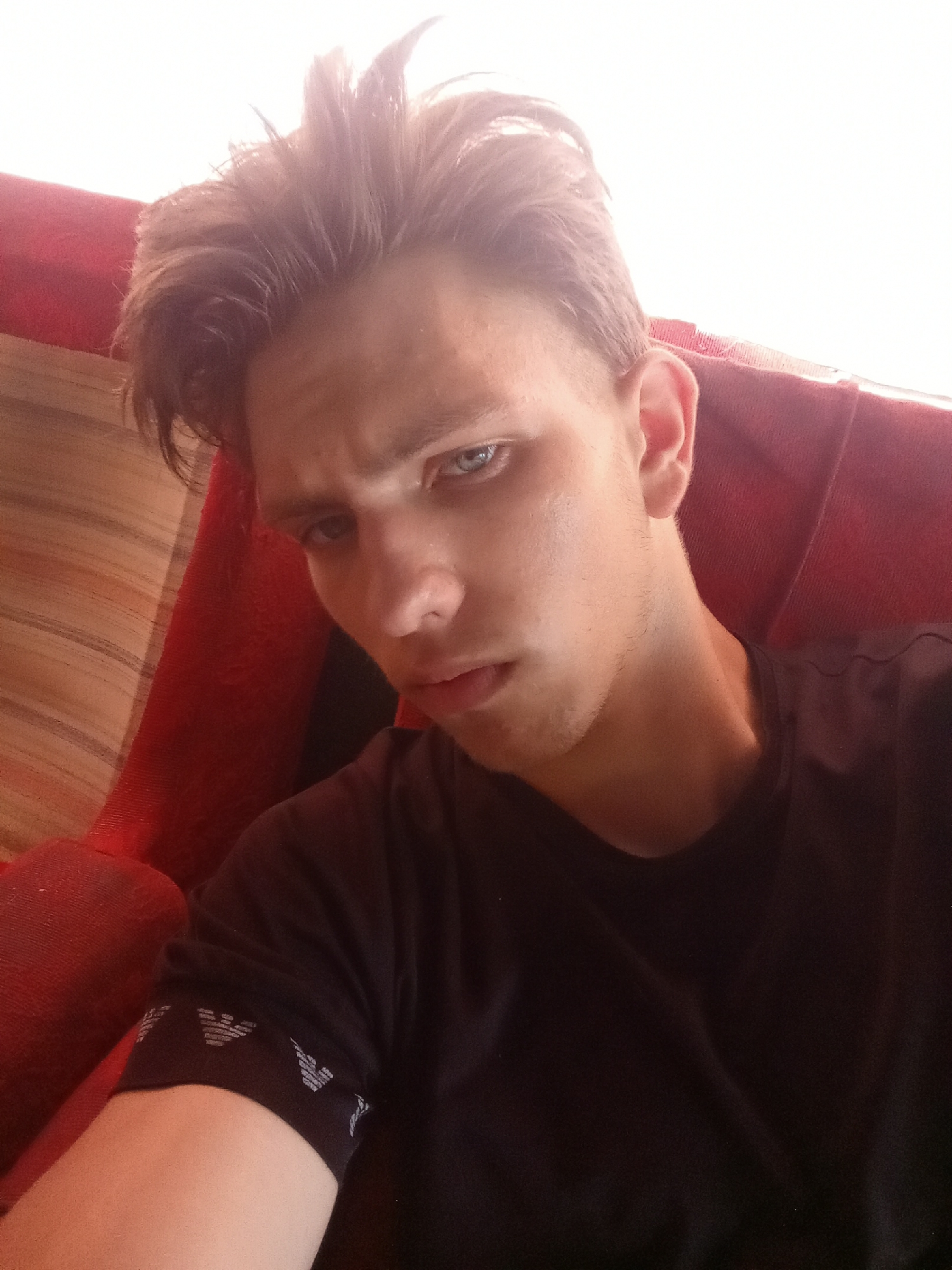 A young man with tousled, light brown hair and blue eyes is pictured in a close-up selfie. He is wearing a black t-shirt with a white patterned band around the sleeve, possibly a brand logo. He is seated on a red upholstered chair, with a wooden-slatted surface visible to the left of the frame. The background is brightly lit, suggesting a daytime setting. The image does not provide any specific information about the city of Zabolotne, Ukraine.