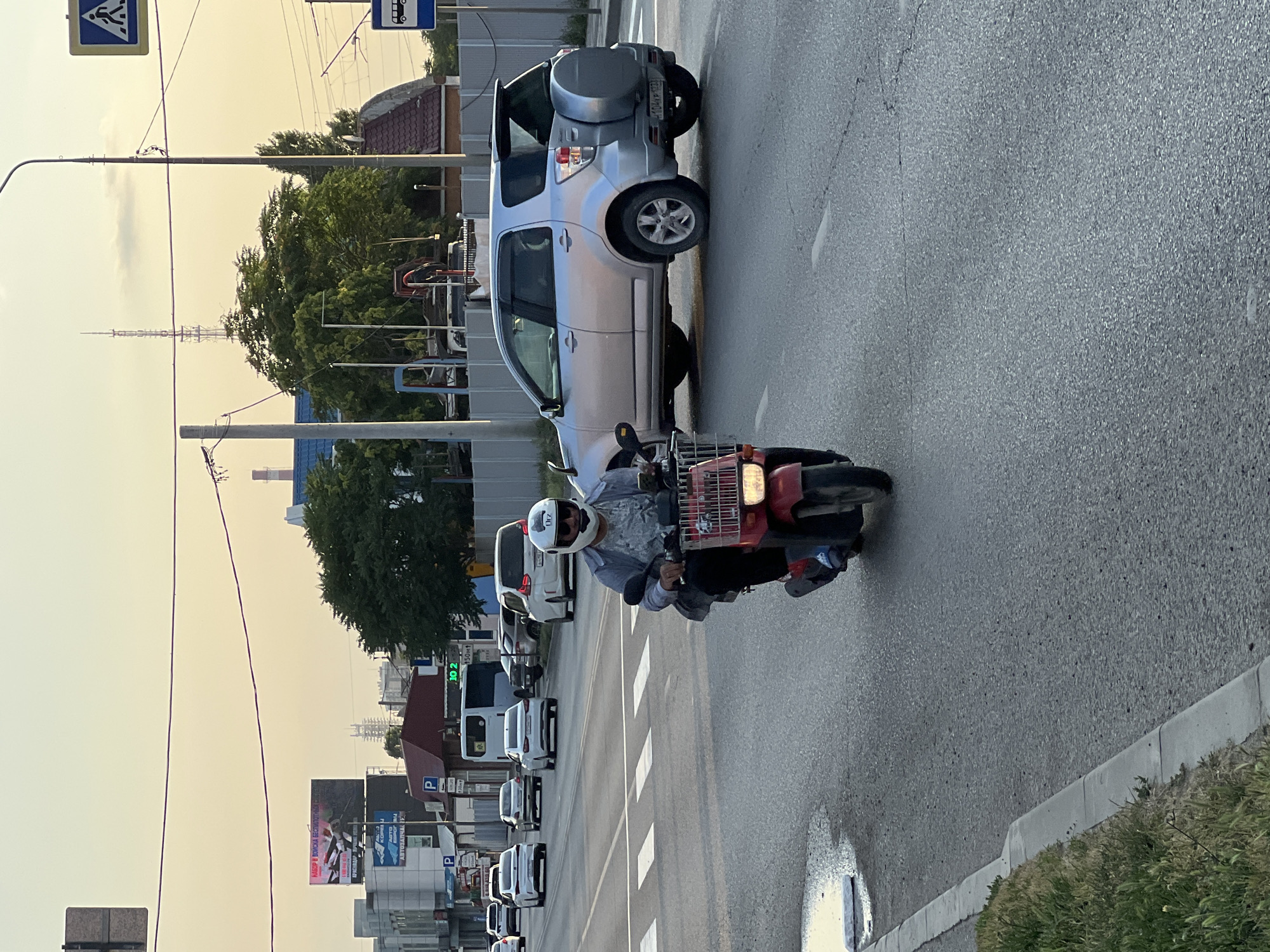 A person rides a scooter on a street in Krasnodar, Russia. The rider is wearing a white helmet and a grey shirt. They are on a red scooter with a white basket. The scooter is in motion, and the rider appears to be looking forward. The scene is a city street with a sidewalk on the right side and a road with multiple lanes. There are cars parked on the left side of the road and some buildings visible in the background. The sky is a pale yellow, suggesting it is either sunrise or sunset. The lighting suggests it is a clear day. There is a pedestrian crossing sign on the left side of the image. The road is wet in some areas, possibly due to recent rain or cleaning. The overall atmosphere appears to be calm and ordinary, capturing a moment of daily life in the city.