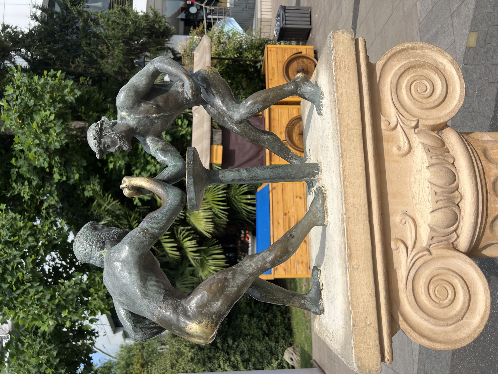 A bronze sculpture depicting two muscular men engaged in an arm wrestling match stands on a stone pedestal. The pedestal is adorned with classical Ionic volutes. The men are intensely focused on their struggle, their bodies strained and muscles bulging. The sculpture is situated outdoors in what appears to be a public space in Sochi, Russia, with lush green trees and foliage in the background. Sunlight illuminates the scene, casting shadows on the sculpture and surrounding environment. Wooden carts with visible wheels are present behind the sculpture, suggesting a marketplace or recreational area. A modern building and a dark trash receptacle are also visible in the background.